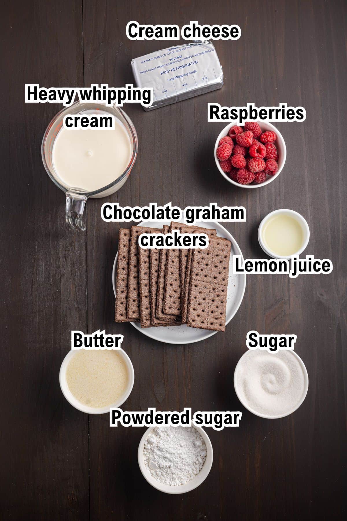 Labeled ingredients for Raspberry Cream Pie on a dark table: cream cheese, heavy whipping cream, raspberries, chocolate graham crackers, lemon juice, butter, sugar, and powdered sugar in separate bowls or plates.