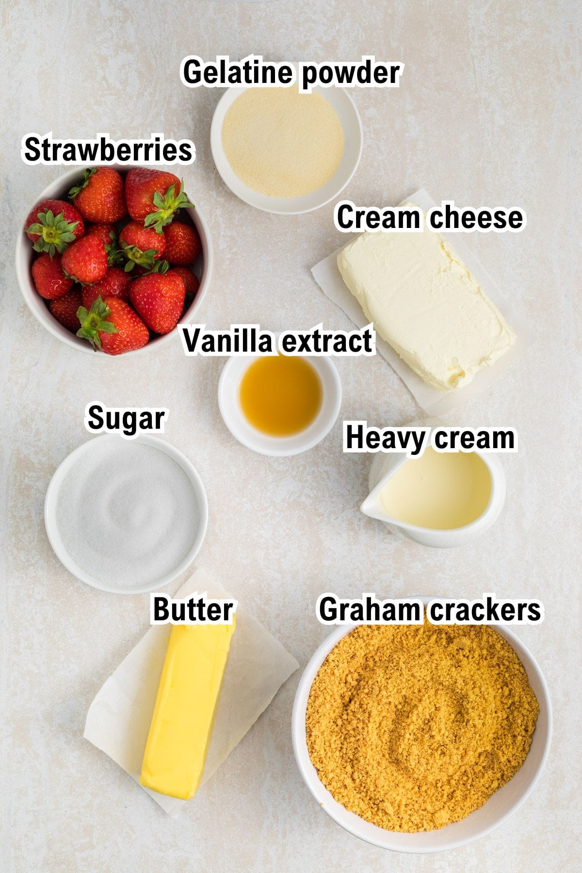 This photo shows the ingredients for No Bake Strawberry Cheesecake: Eight labeled ingredients are arranged on a light surface: strawberries, gelatine powder, cream cheese, vanilla extract, heavy cream, sugar, butter, and graham cracker crumbs. Each ingredient is in a bowl or on a plate.