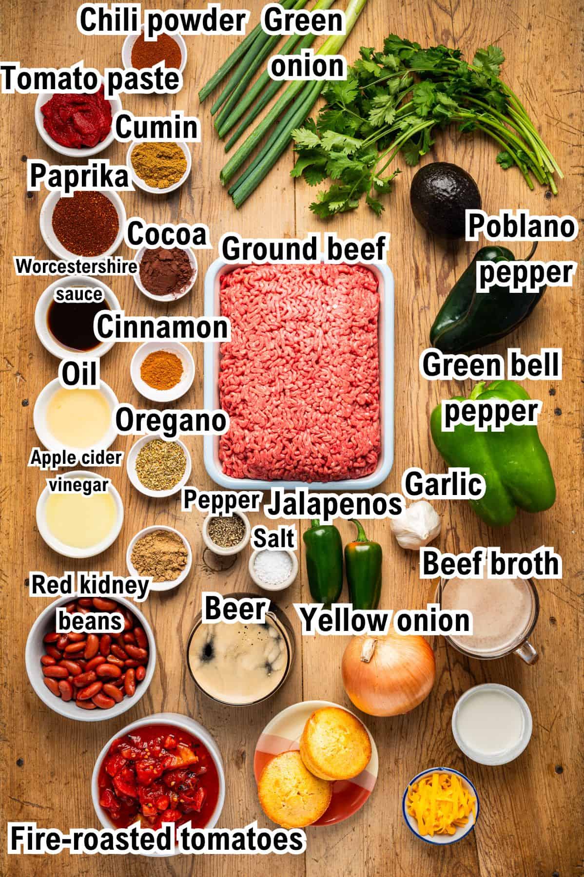 The Ingredients for Beef Chili Top-down view of labeled beef chili ingredients, including ground beef, beans, vegetables, spices, beer, broth, and herbs, all neatly arranged on a wooden surface.
