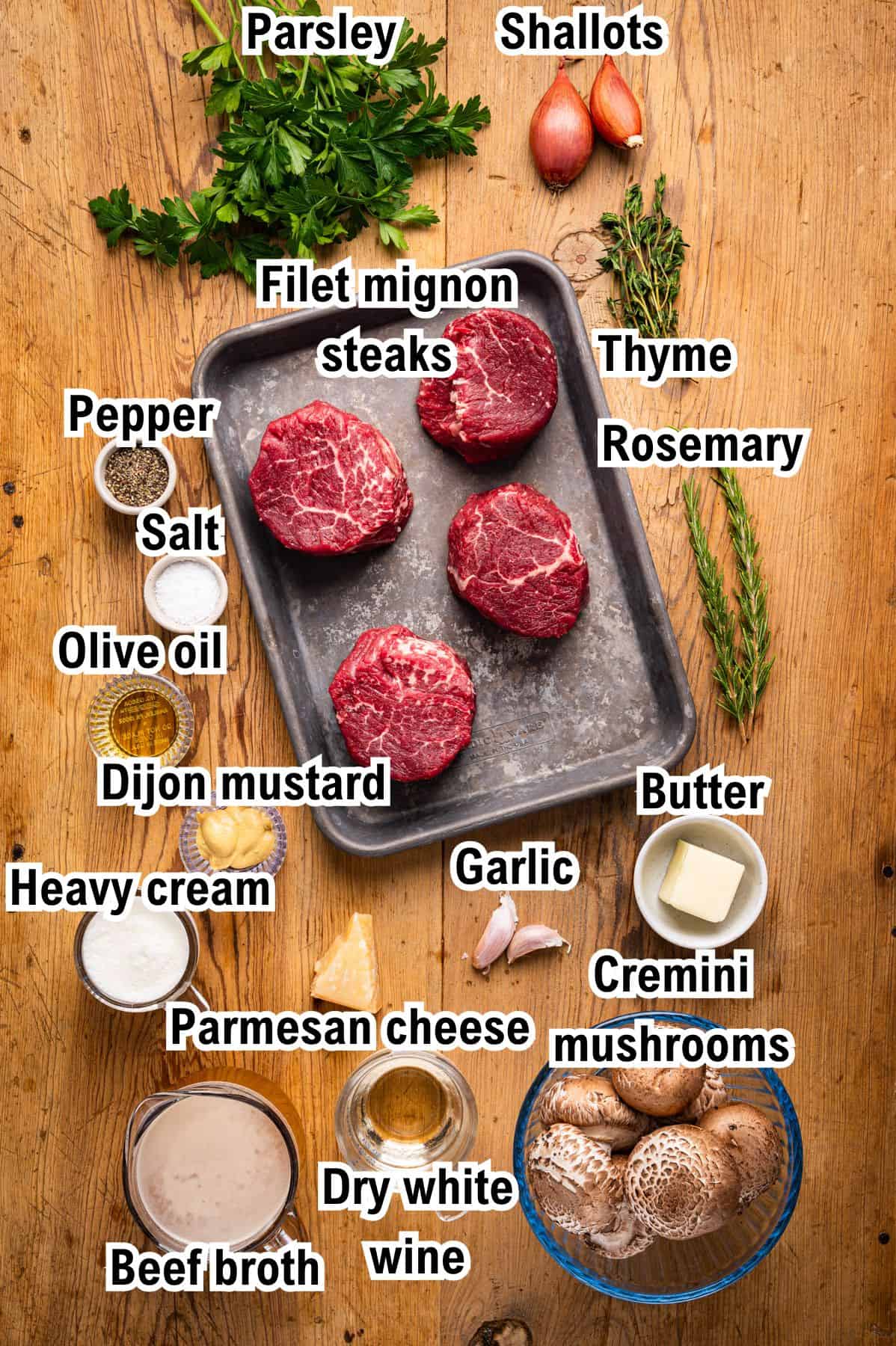 A wooden table with labeled ingredients for filet mignon: parsley, shallots, thyme, rosemary, filet mignon steaks, pepper, salt, olive oil, Dijon mustard, heavy cream, Parmesan cheese, beef broth, dry white wine, garlic, butter, and cremini mushrooms.