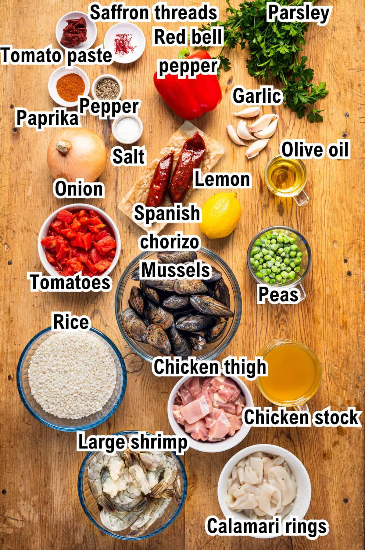 The Ingredients for Spanish Paella Top-down view of paella ingredients, including labeled bowls of rice, tomatoes, peas, chorizo, mussels, chicken, shrimp, calamari, spices, herbs, olive oil, lemon, and a red bell pepper.