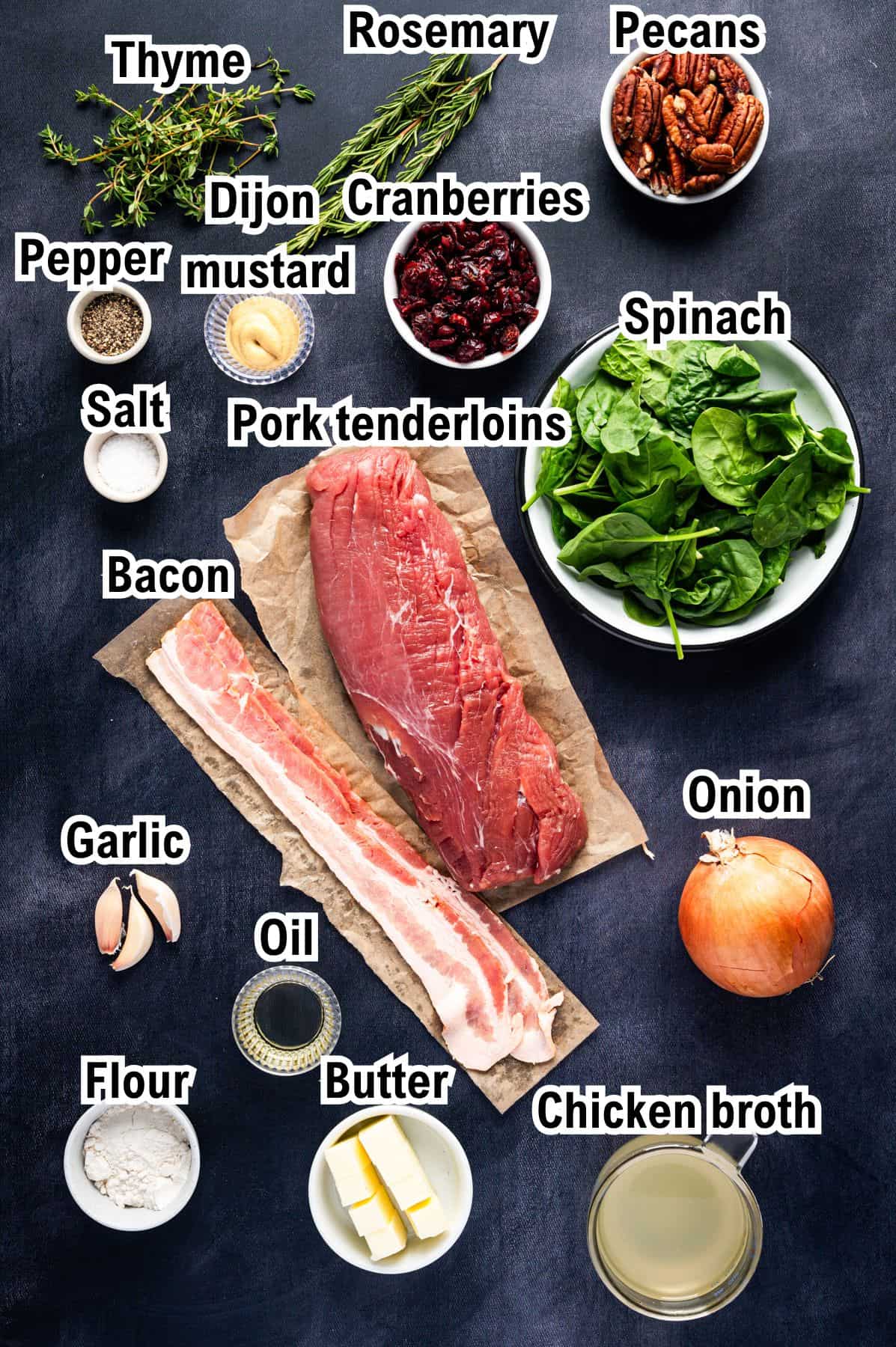 The Ingredients for Stuffed Pork Tenderloin A flat lay of labeled ingredients on a dark surface, including pork tenderloin, bacon, spinach, pecans, cranberries, onion, garlic, salt, pepper, thyme, rosemary, Dijon mustard, flour, butter, oil, and chicken broth.