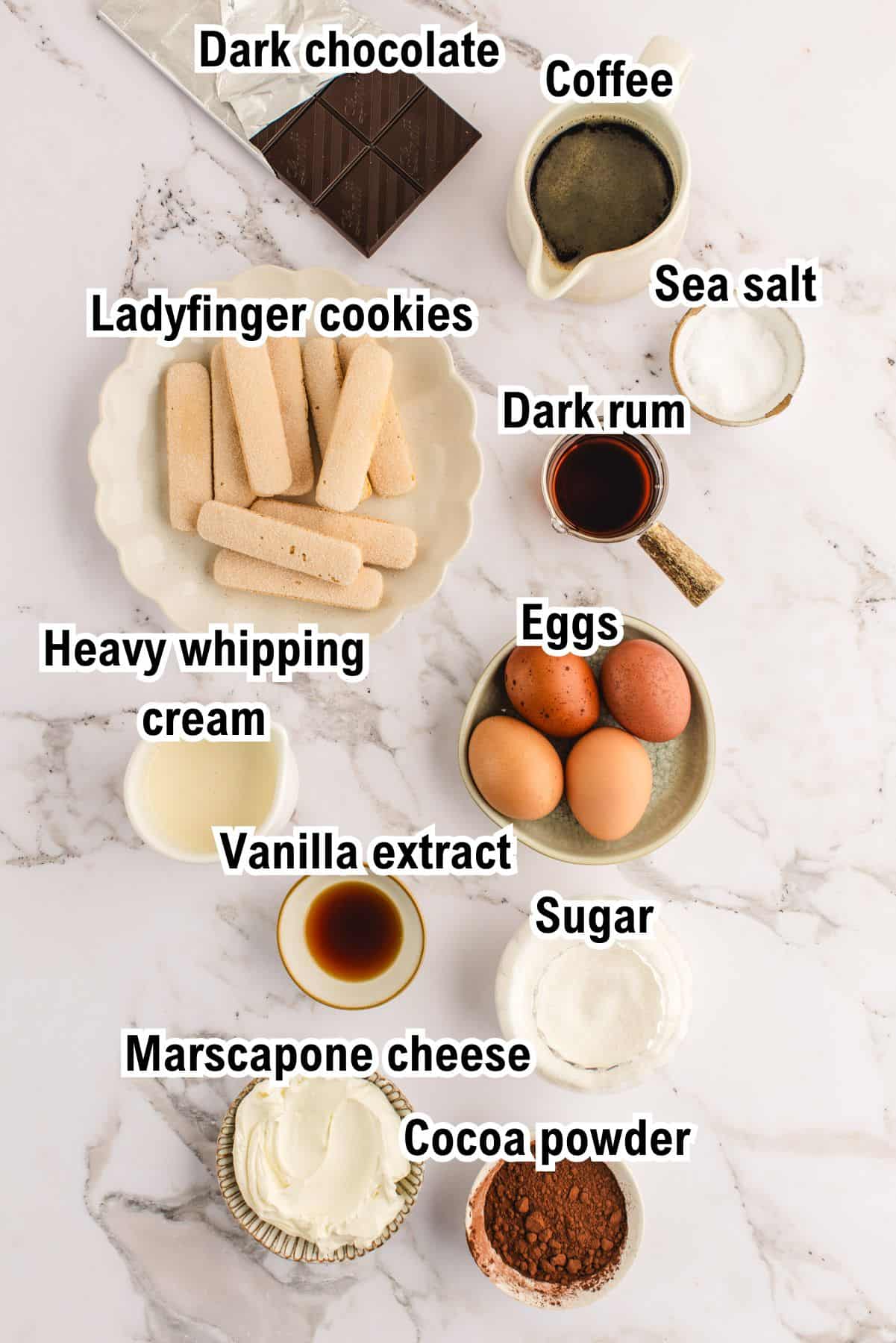 The Ingredients for Tiramisu Labeled ingredients for tiramisu arranged on a marble surface: dark chocolate, coffee, sea salt, ladyfinger cookies, dark rum, eggs, heavy whipping cream, vanilla extract, sugar, mascarpone cheese, and cocoa powder.