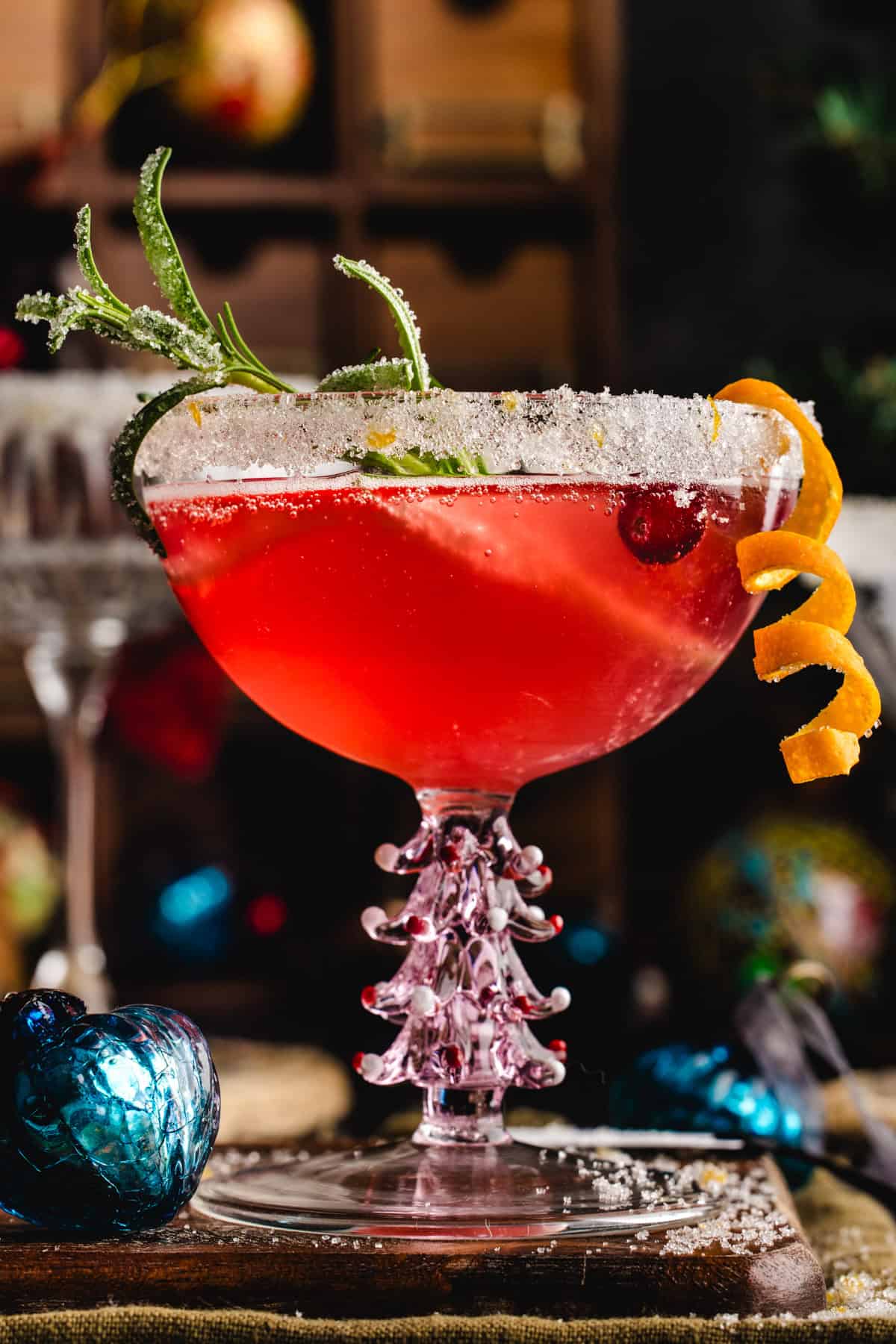 Christmas Mimosa (red in color from cranberry juice) in a glass, garnished with sugared rosemary and an orange peel twist.