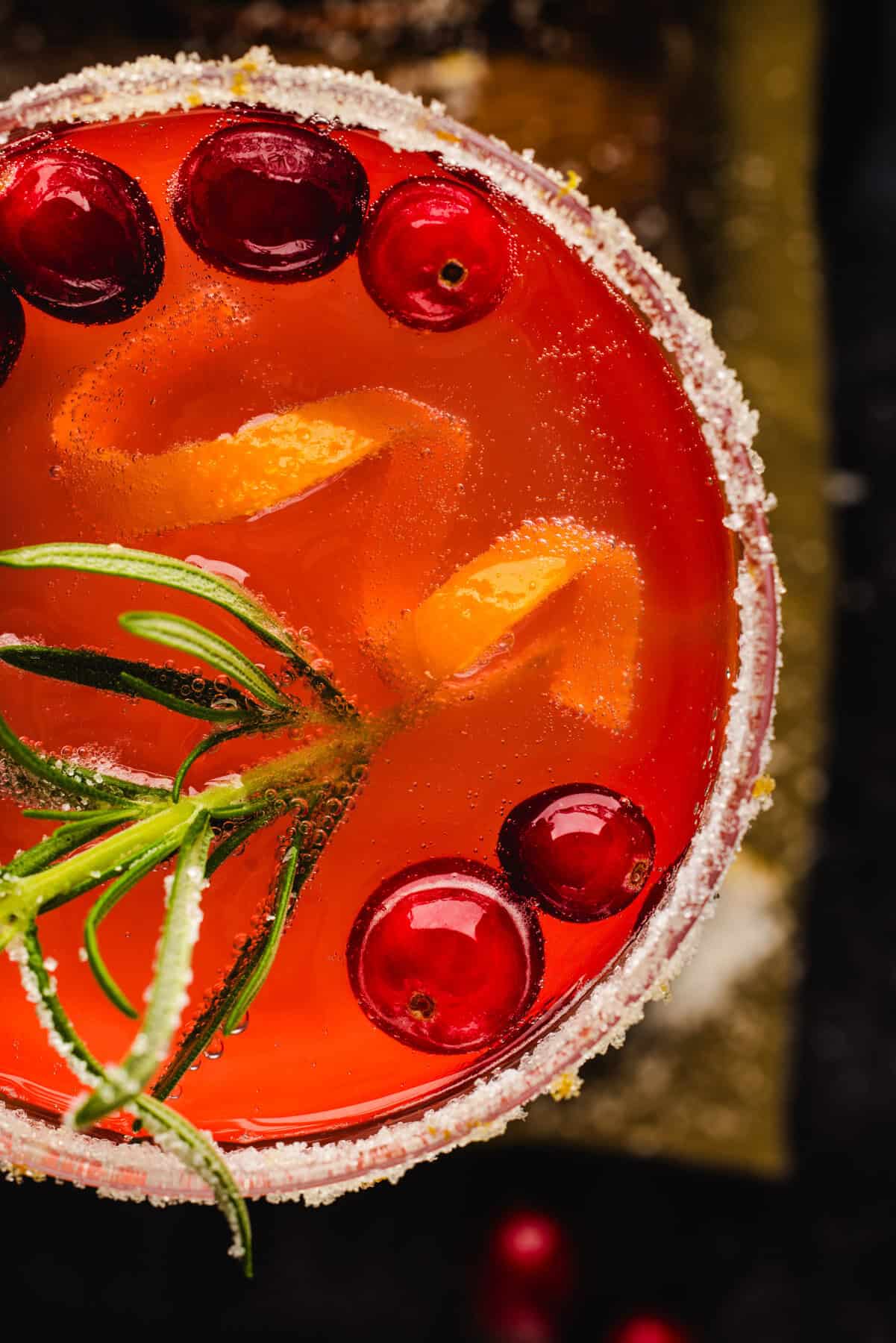 Overhead close-up shot of Christmas Mimosa (red in color from cranberry juice) in a glass, garnished with sugared rosemary, fresh cranberries, and an orange peel twist.