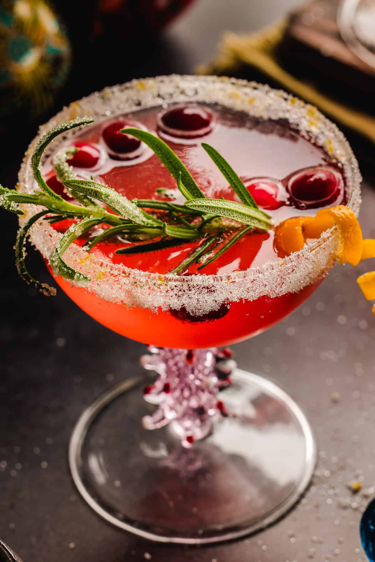 Christmas Mimosa (red in color from cranberry juice) in a glass, garnished with sugared rosemary, fresh cranberries, and an orange peel twist.