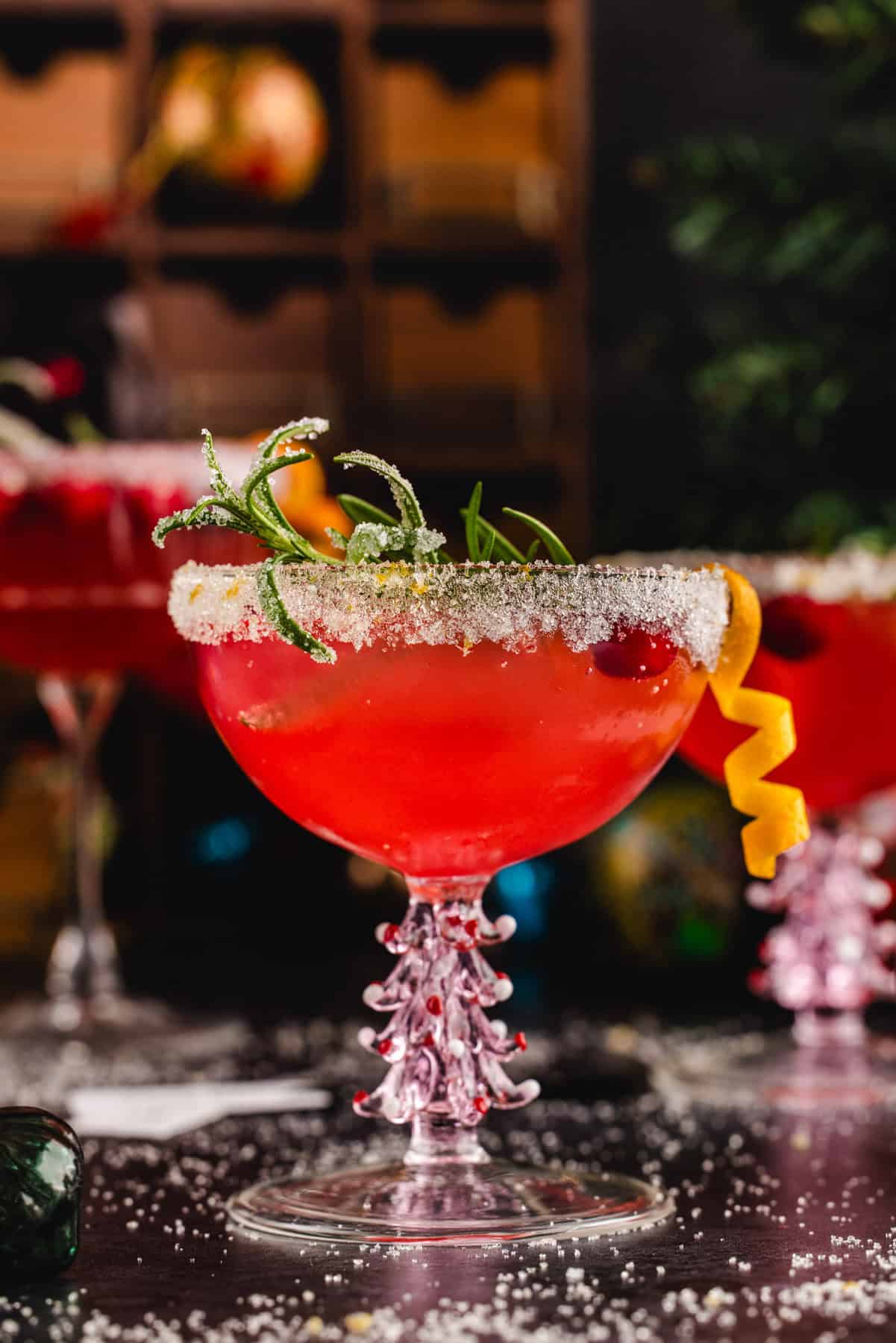 Christmas Mimosa (red in color from cranberry juice) in a glass, garnished with sugared rosemary, fresh cranberries, and an orange peel twist.