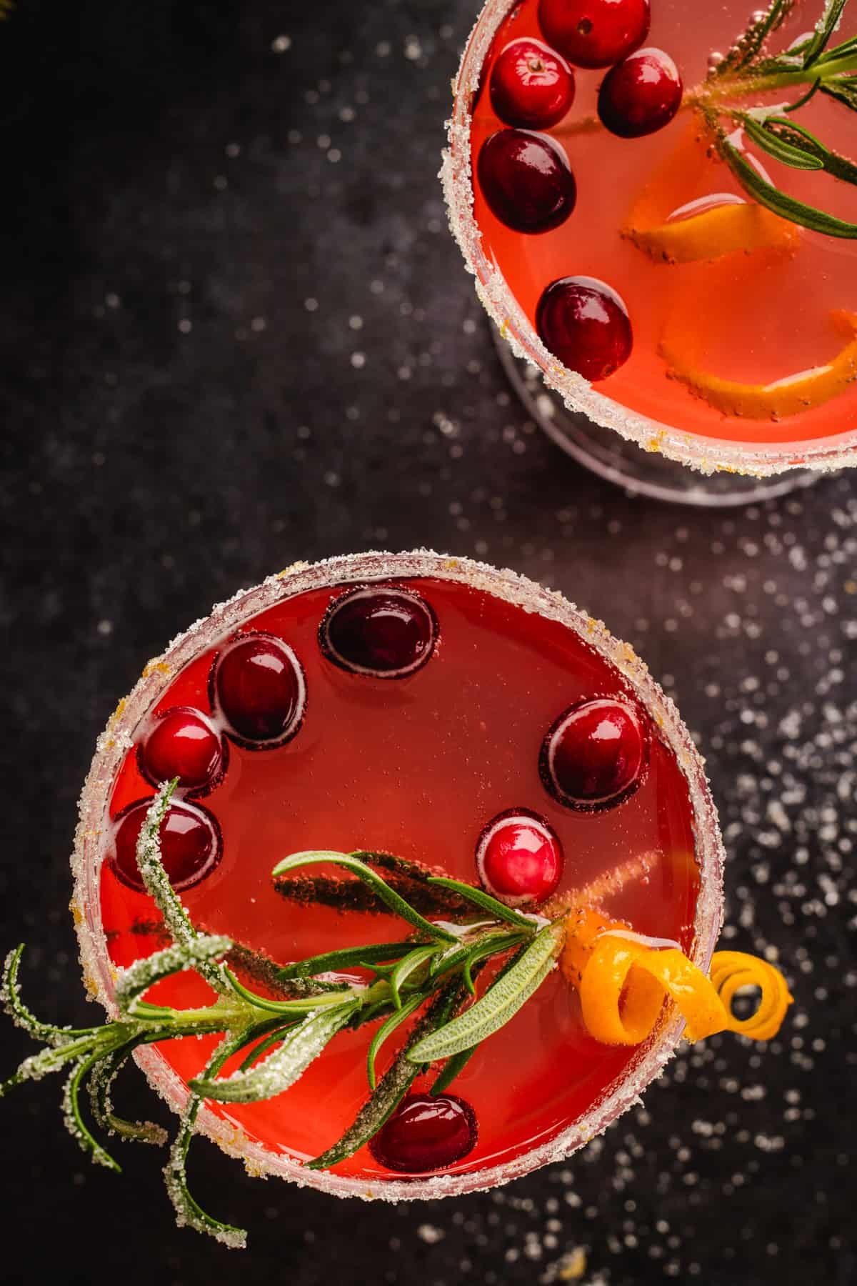 Overhead view of Christmas Mimosa (red in color from cranberry juice) in 2 glasses, garnished with sugared rosemary, fresh cranberries, and an orange peel twist.
