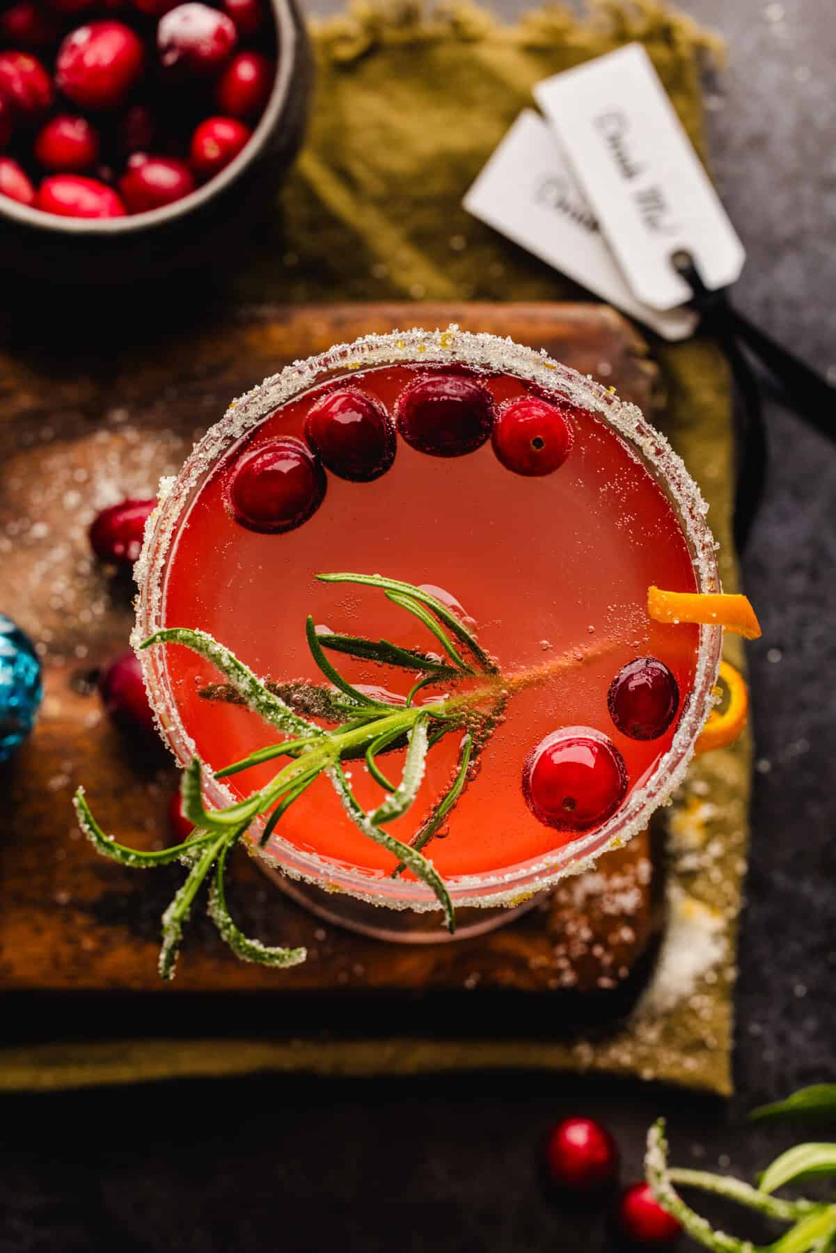The overhead view of a Christmas Mimosa (red from cranberry juice) in a glass, garnished with sugared rosemary, fresh cranberries, and an orange peel twist.