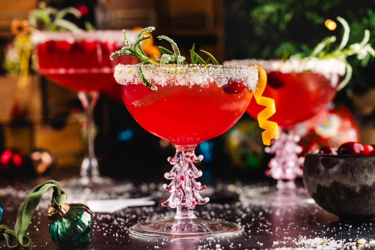3 Christmas Mimosa glasses (red in color from cranberry juice), garnished with sugared rosemary, fresh cranberries, and an orange peel twist.