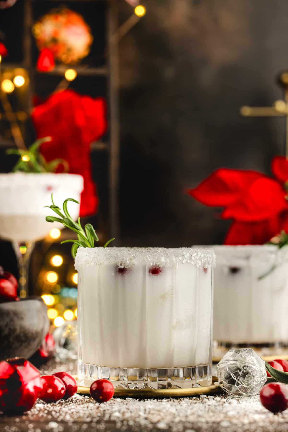 White Christmas Margarita in a glass, garnished with fresh cranberries and a rosemary sprig - against a dark festive background with bright red ribbons in the back.