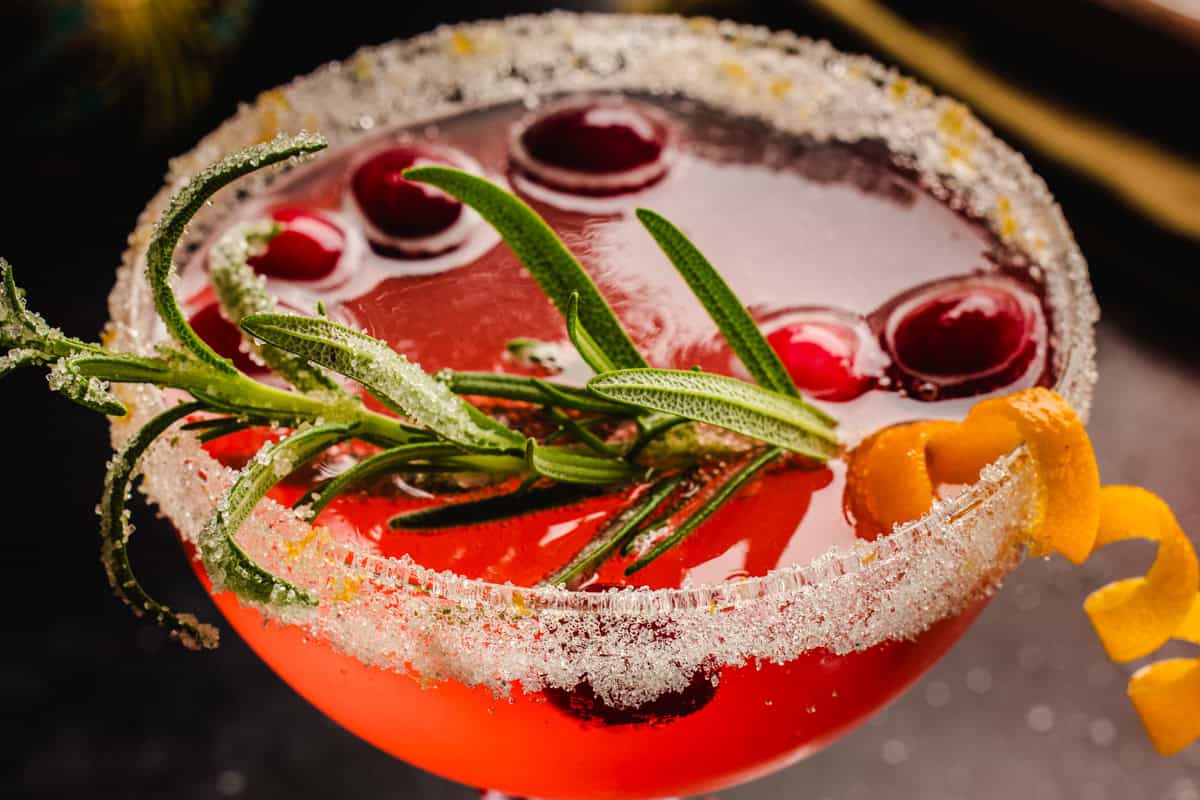 Christmas Mimosa (red in color from cranberry juice) in a glass, garnished with sugared rosemary, fresh cranberries, and an orange peel twist.