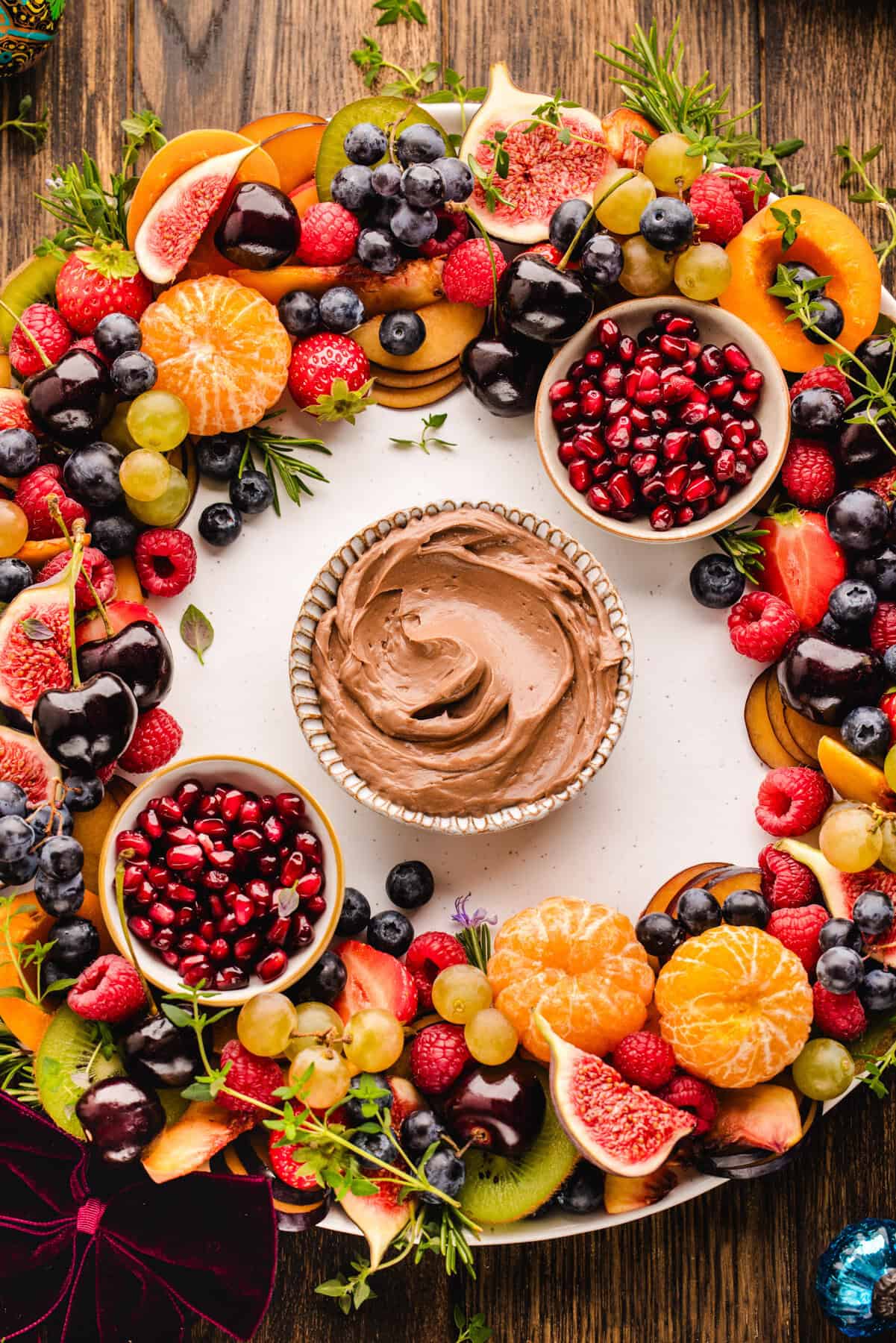Fruit Platter with Nutella Dip A round fruit wreath arranged on a large platter, with colorful sliced fruit layered in a circular pattern and two small bowls of pomegranate arils tucked on opposite sides, with Nutella dip in a bowl in the center of the wreath.
