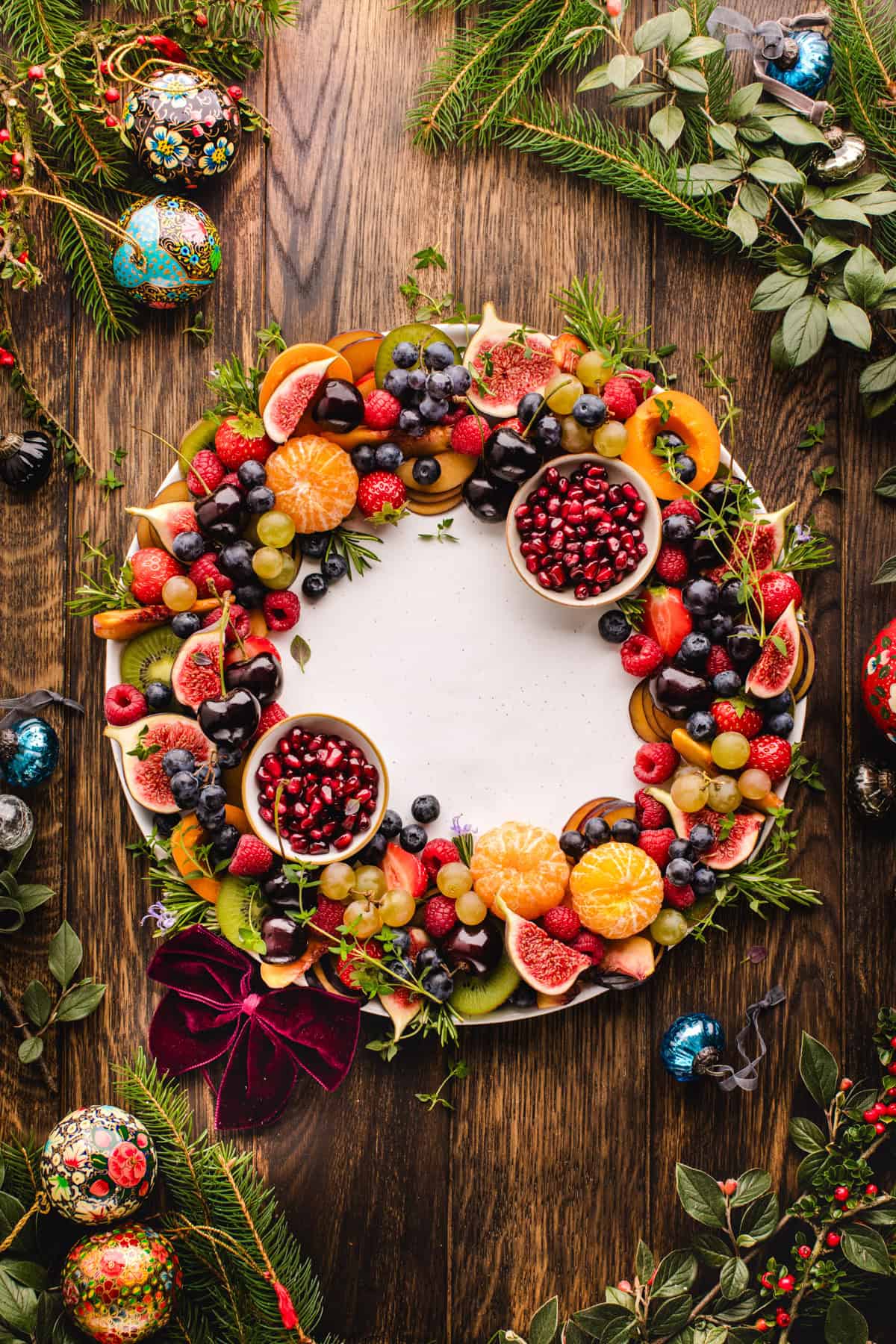 Fruit Platter A round fruit wreath arranged on a large platter, with colorful sliced fruit layered in a circular pattern and two small bowls of pomegranate arils tucked on opposite sides, with Nutella dip in a bowl in the center of the wreath.
