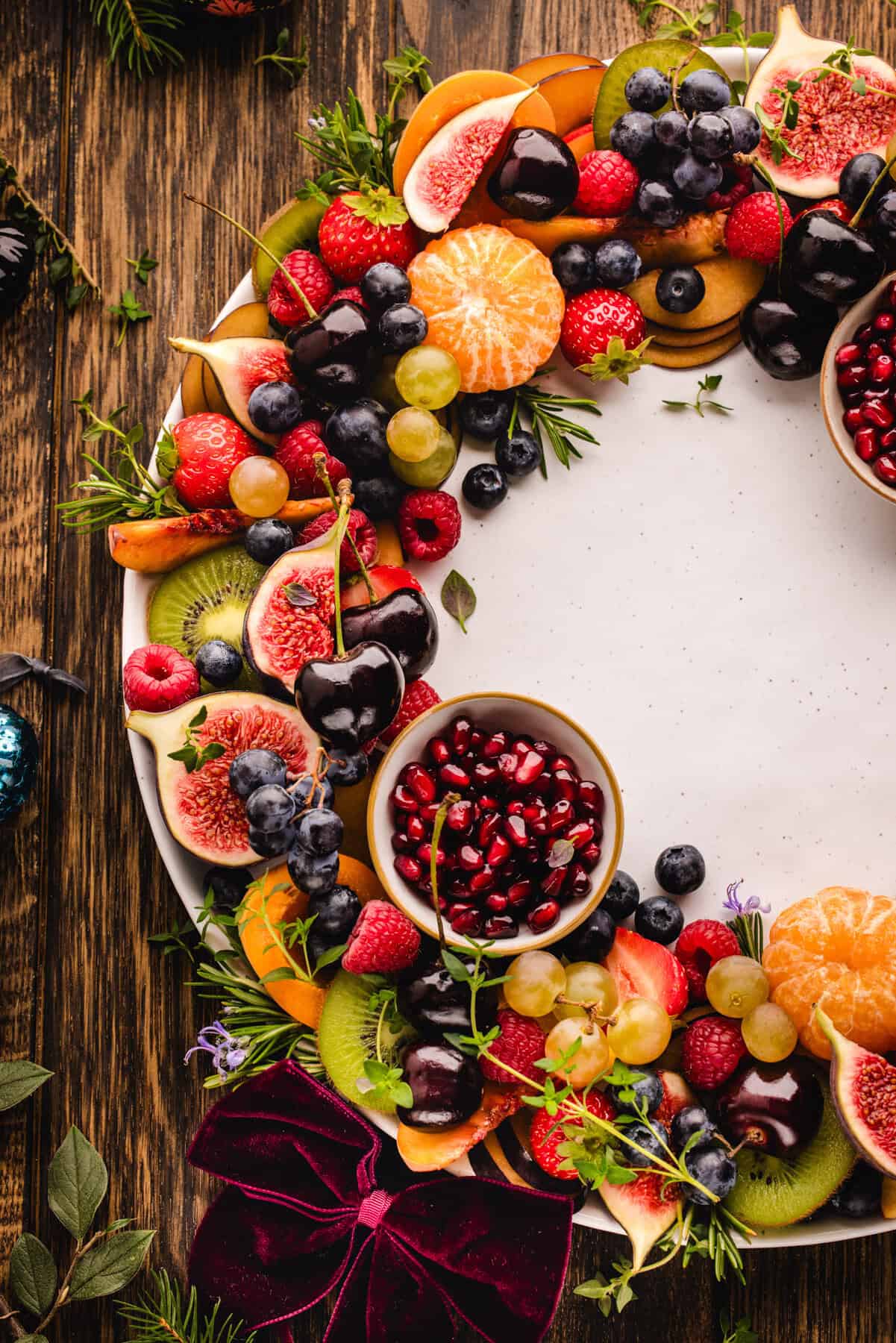 Fruit Platter A round fruit wreath arranged on a large platter, with colorful sliced fruit layered in a circular pattern and two small bowls of pomegranate arils tucked on opposite sides.