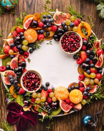 A round fruit wreath arranged on a large platter, with colorful sliced fruit layered in a circular pattern and two small bowls of pomegranate arils tucked on opposite sides.