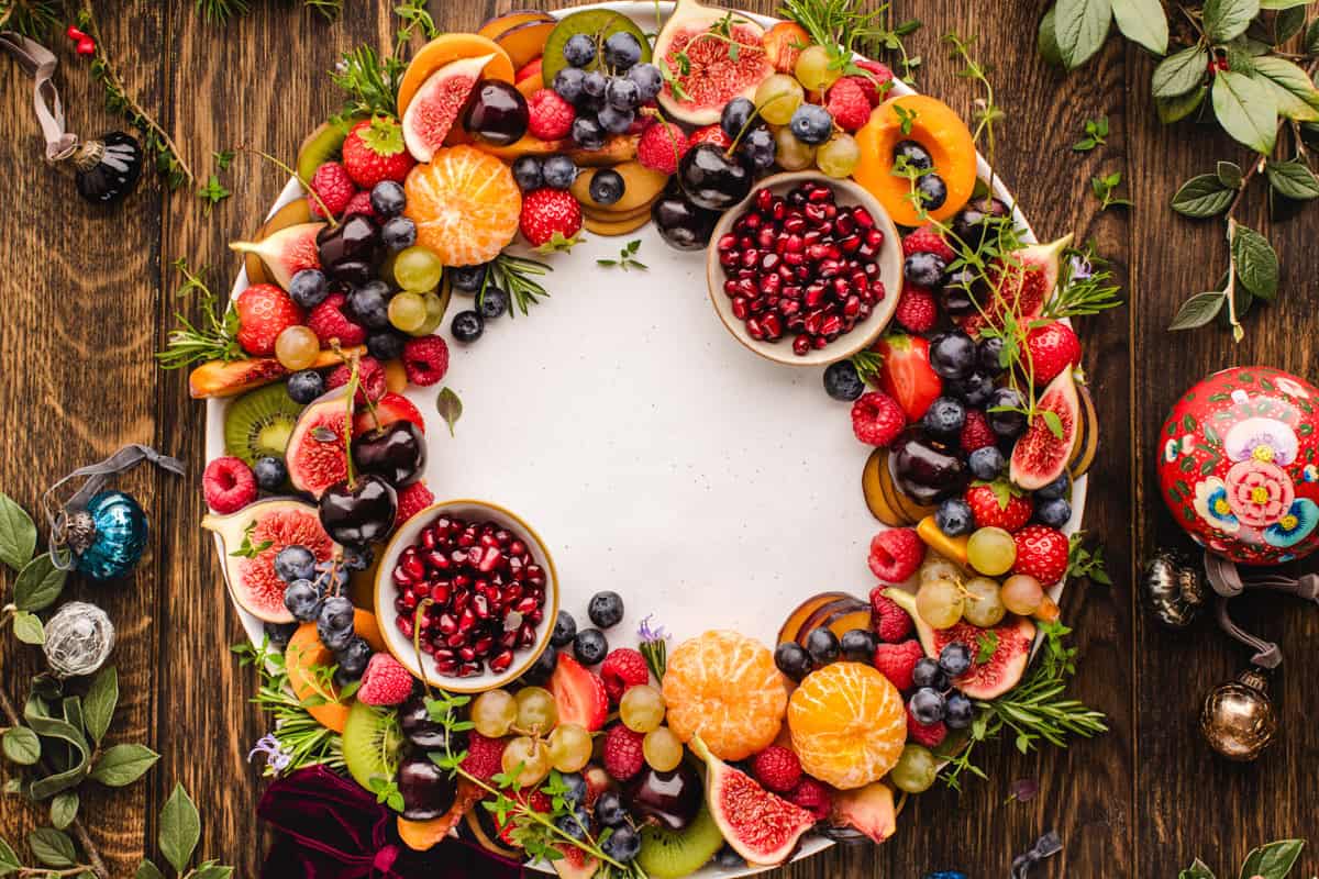 Fruit Platter A round fruit wreath arranged on a large platter, with colorful sliced fruit layered in a circular pattern and two small bowls of pomegranate arils tucked on opposite sides.