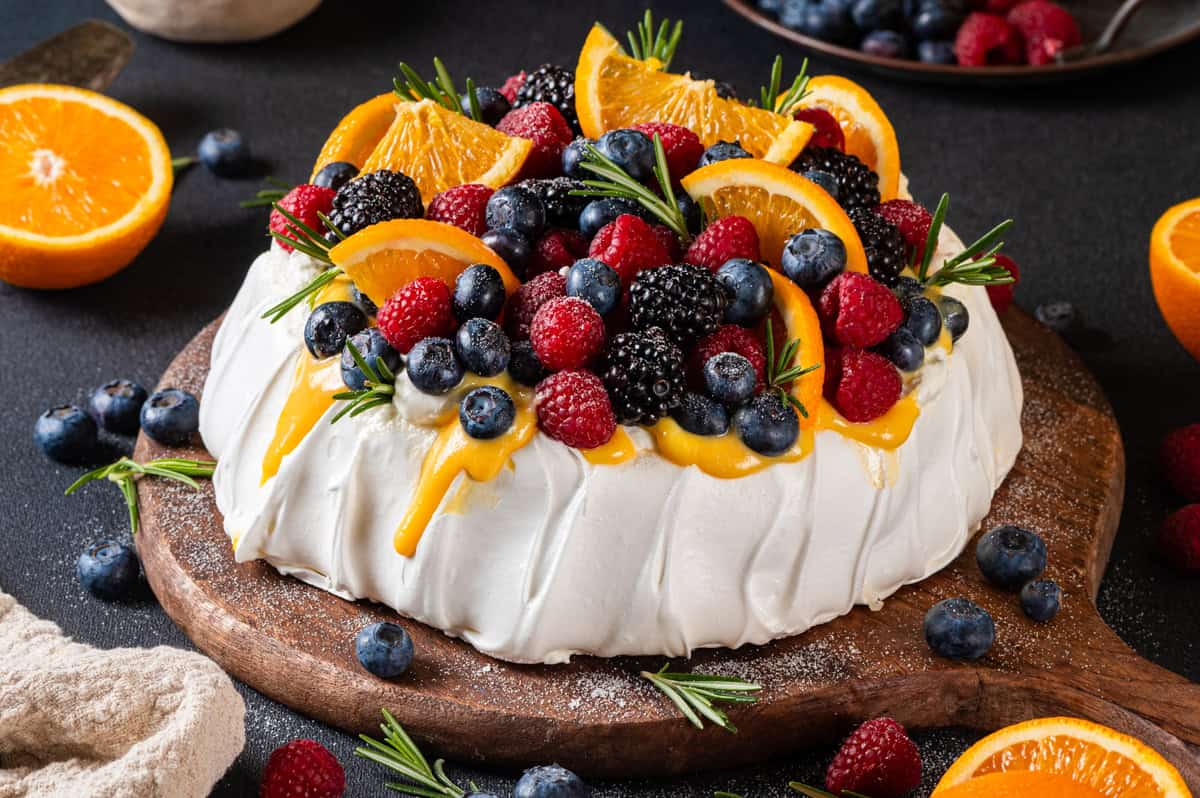 Classic Pavolva Dessert A pavlova dessert topped with raspberries, blueberries, blackberries, orange slices, and sprigs of rosemary sits on a wooden board, surrounded by more berries and halved oranges on a dark surface.