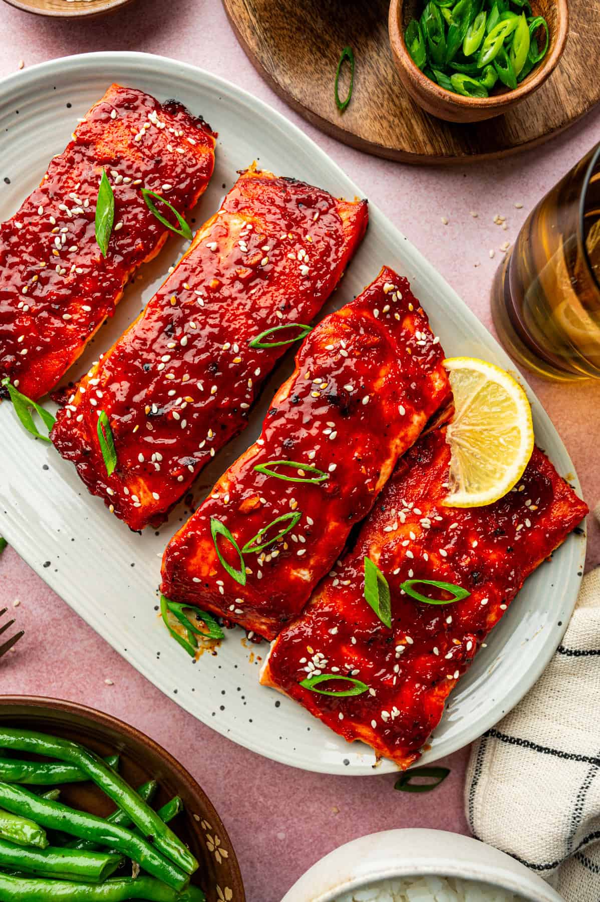 A plate with four gochujang salmon fillets glazed to perfection, garnished with sesame seeds and sliced green onions, served with a lemon wedge. Side dishes of green beans, white rice, and a drink are visible nearby.