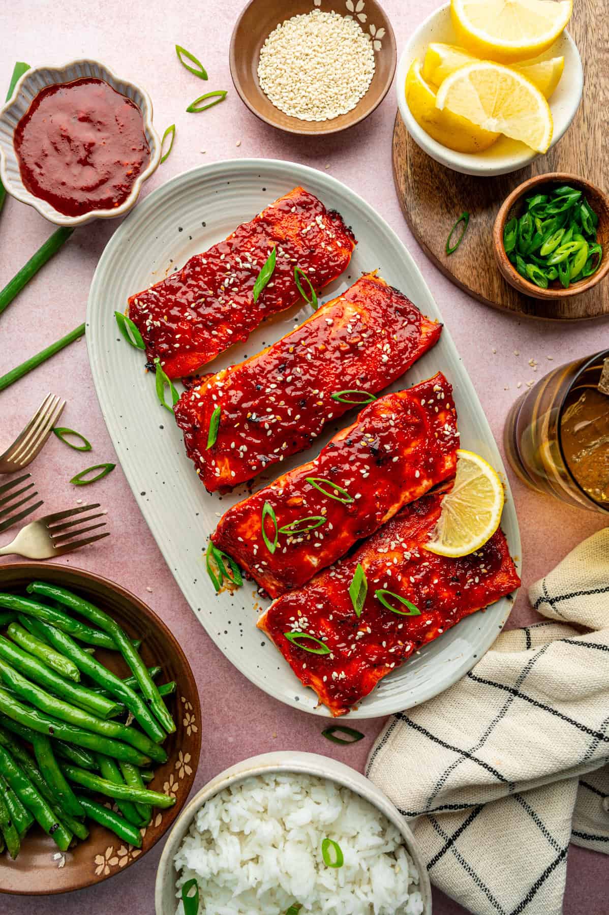 An overhead view of a plate with four gochujang salmon fillets glazed and garnished with sesame seeds and green onions, surrounded by lemon wedges, green beans, white rice, sauce, sesame seeds, and sliced green onions.