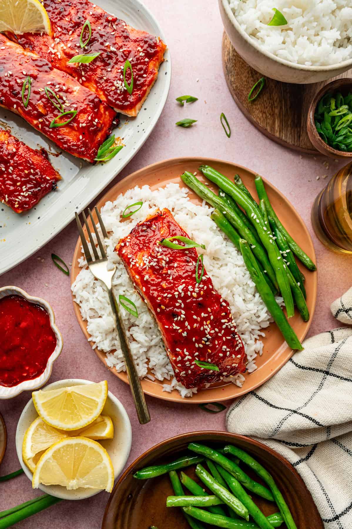 A plate with white rice, green beans, and gochujang salmon fillet topped with sesame seeds and sliced green onions. Surrounding the plate are extra salmon, rice, green beans, sauce, lemon wedges, and a napkin.