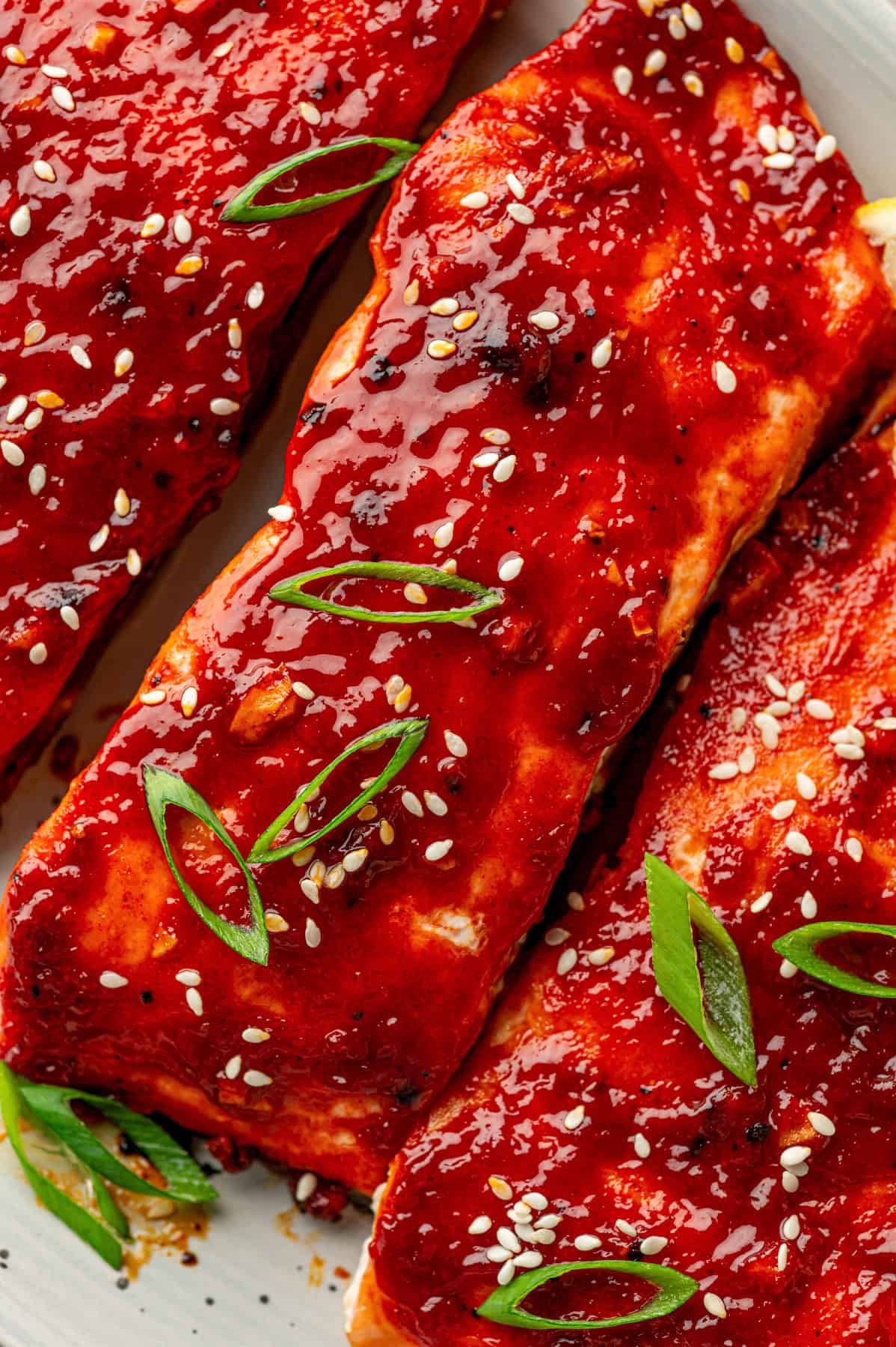 Close-up of gochujang salmon fillets glazed with a shiny red sauce, garnished with sesame seeds and sliced green onions, served on a white plate.