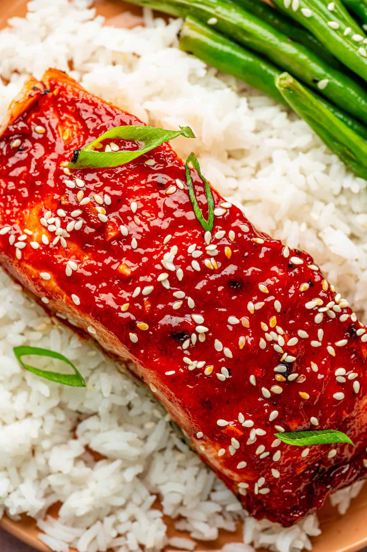 A fillet of gochujang salmon glazed and topped with sesame seeds and sliced green onions, served on a bed of white rice with green beans on the side.