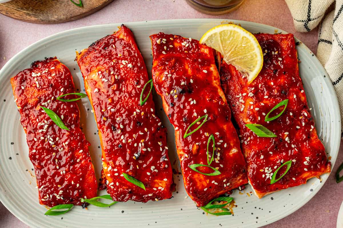 Four pieces of gochujang salmon, glazed and topped with sesame seeds and sliced green onions, are arranged on a white oval plate and garnished with a lemon wedge.