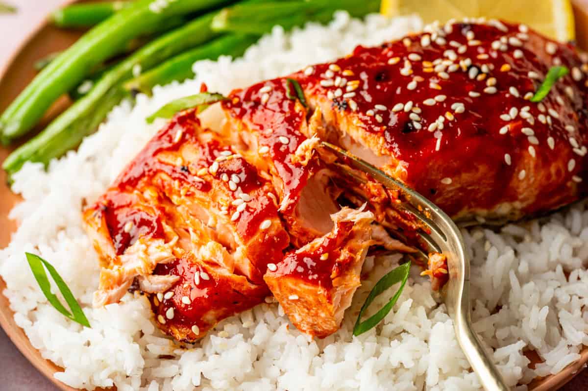A fork flakes a piece of gochujang salmon glazed and topped with sesame seeds and green onions, served on a bed of white rice with green beans and a lemon wedge on the side.