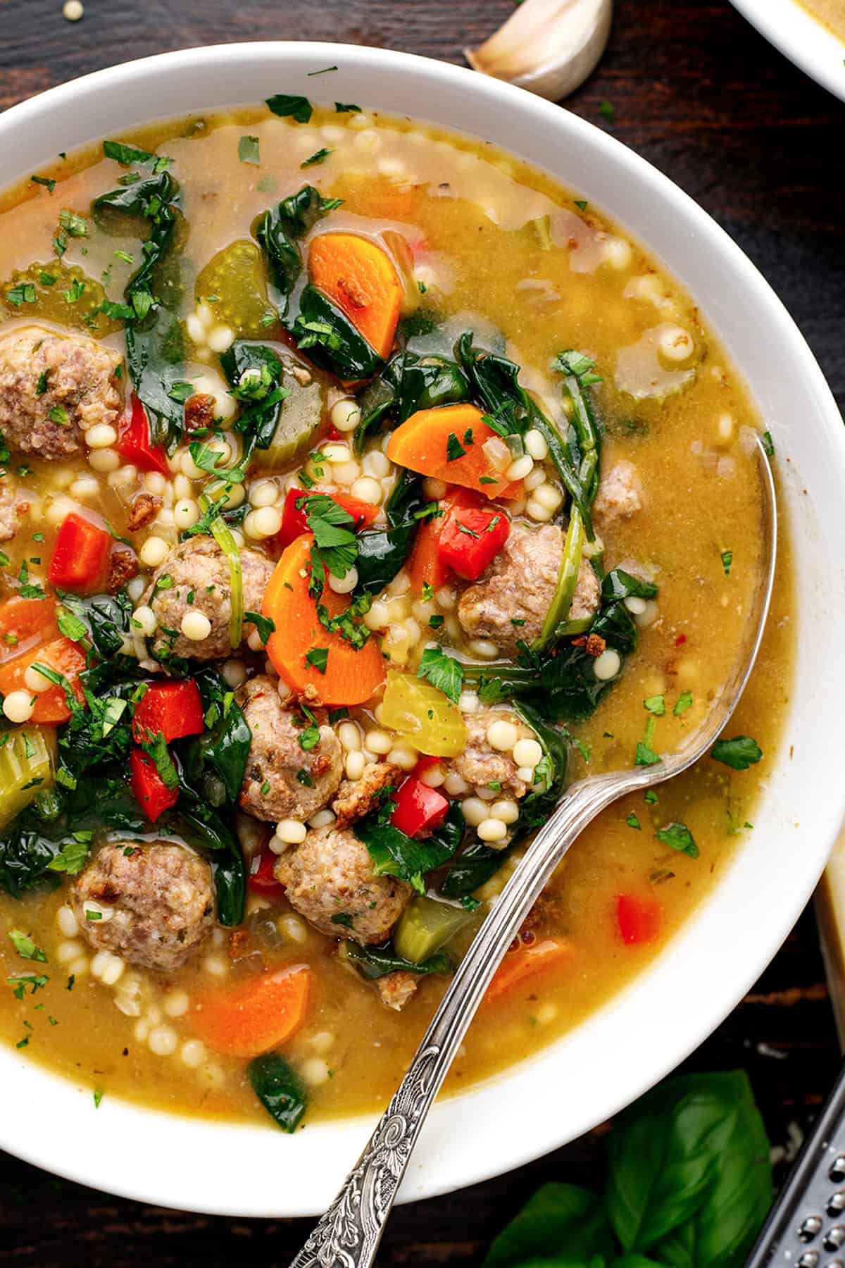 Italian Wedding Soup A close-up of a bowl of Italian wedding soup filled with meatballs, carrots, spinach, celery, red bell peppers, small pasta (Acini di Pepe), and herbs, with a spoon resting in the bowl.