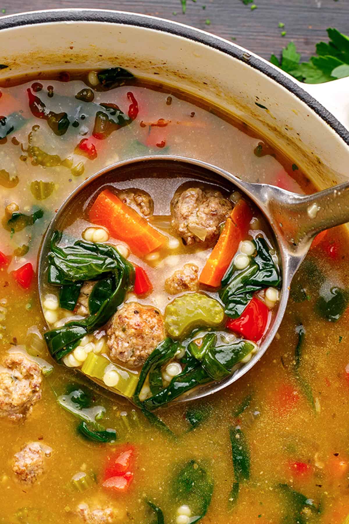 Italian Wedding Soup A ladle lifting a serving of hearty Italian wedding soup with meatballs, carrots, celery, spinach, and small pasta (Acini di Pepe) from a pot, showing colorful vegetables and broth.