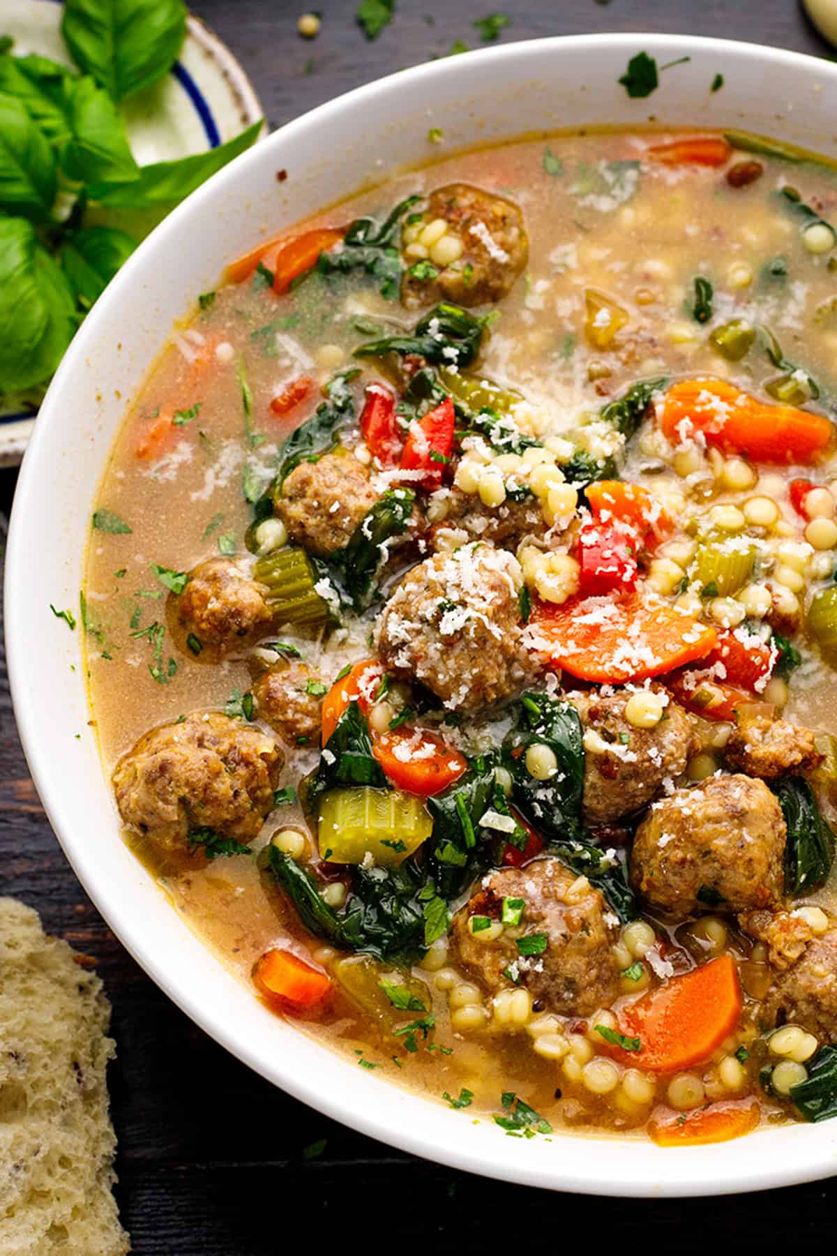 Italian Wedding Soup A close-up of a bowl of Italian wedding soup with meatballs, diced carrots, celery, red bell peppers, greens, small pasta (Acini di Pepe), and sprinkled grated cheese on top. A piece of bread and fresh basil are beside the bowl.