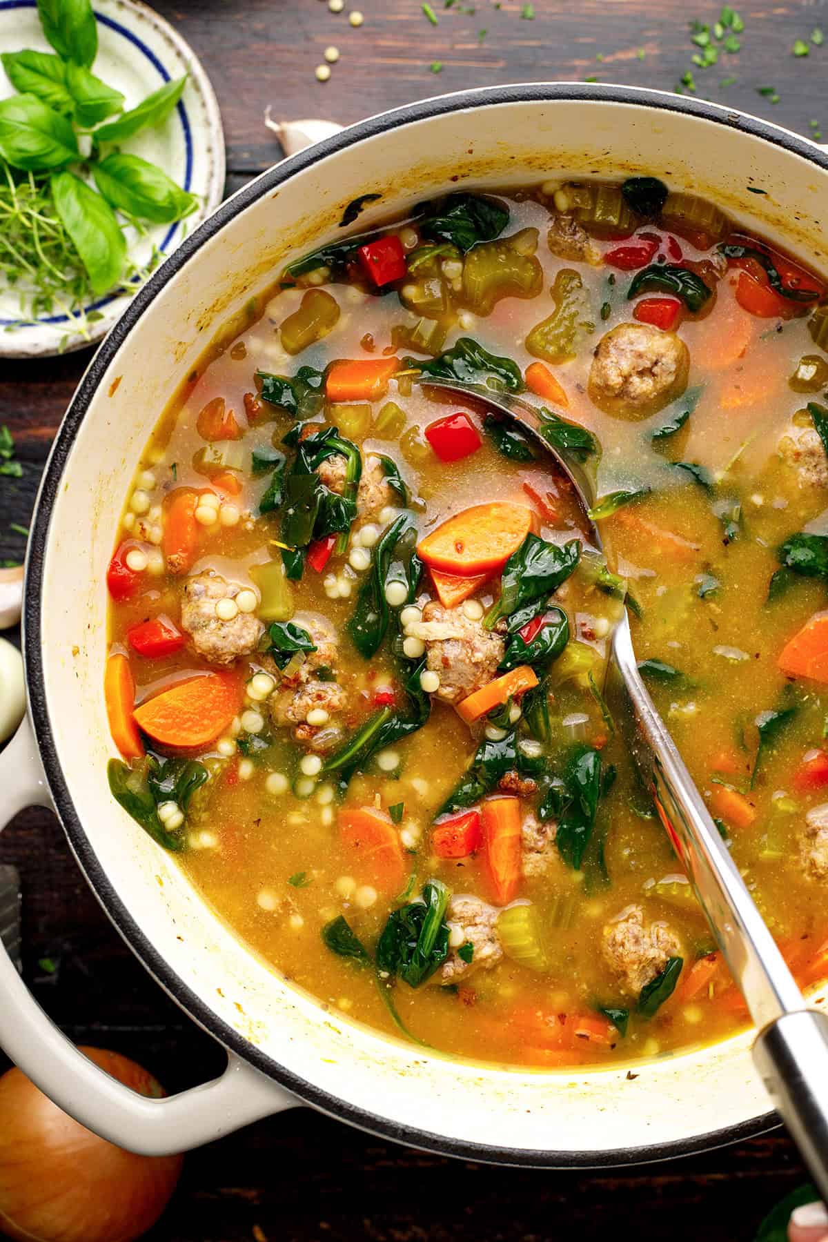 Italian Wedding Soup A pot of homemade Italian wedding soup with meatballs, carrot slices, celery, red bell peppers, leafy greens, and small pasta (Acini di Pepe) in a clear broth, with a ladle inside. Fresh herbs and vegetables are on the table nearby.