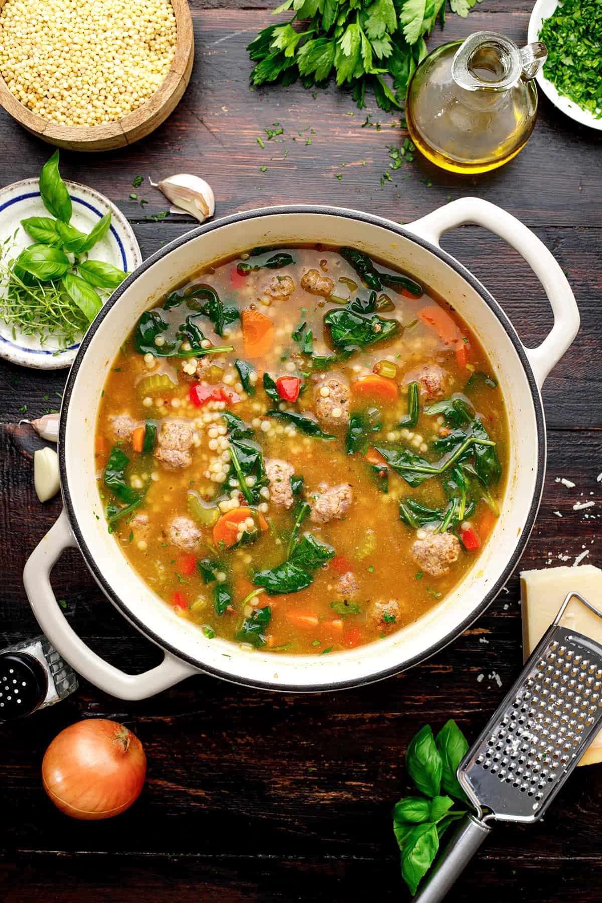 Italian Wedding Soup A pot of Italian wedding soup with meatballs, spinach, carrots, acini di pepe pasta, and broth sits on a wooden table, surrounded by fresh herbs, cheese, olive oil, and vegetables.