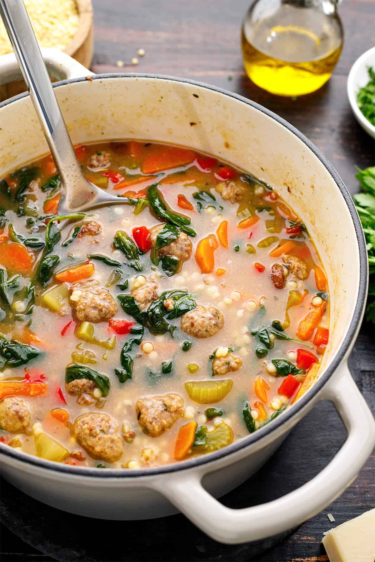 Italian Wedding Soup A white pot filled with Italian wedding soup containing sausage, spinach, carrots, celery, and small pasta (Acini di Pepe), with a metal ladle inside. Olive oil, herbs, and grated cheese are visible in the background on a wooden table.
