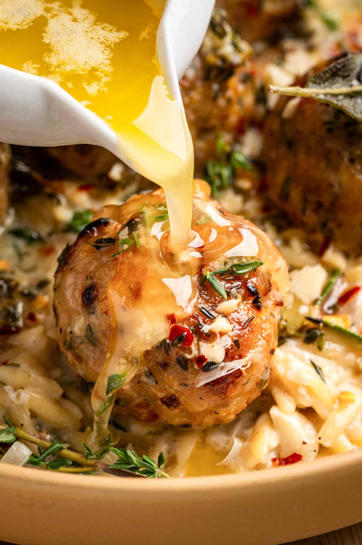 Pouring the sauce over the single chicken meatball nestled in creamy orzo on a plate.