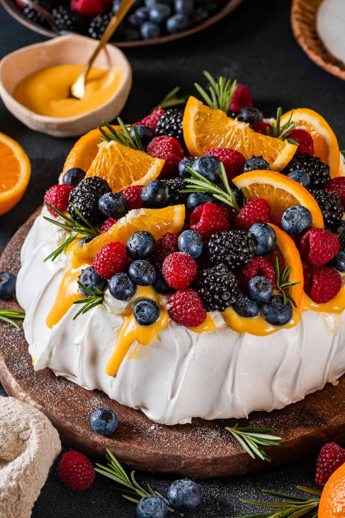 Classic Pavolva Dessert A pavlova dessert topped with fresh blueberries, raspberries, blackberries, orange slices, rosemary sprigs, and a drizzle of yellow sauce (orange curd) sits on a wooden board.