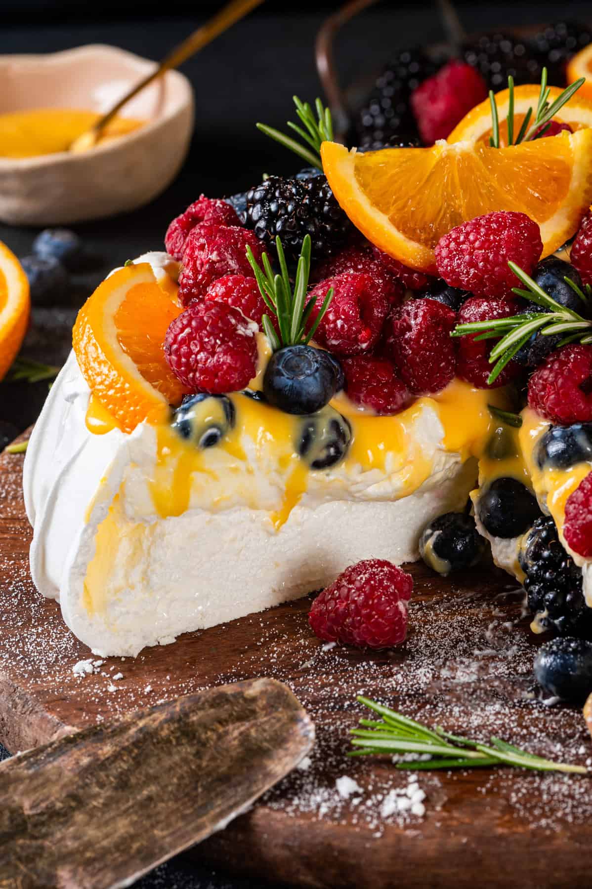 Classic Pavolva Dessert A pavlova dessert topped with fresh raspberries, blueberries, blackberries, orange slices, rosemary sprigs, and orange curd sauce sits on a wooden board with a slice cut out.