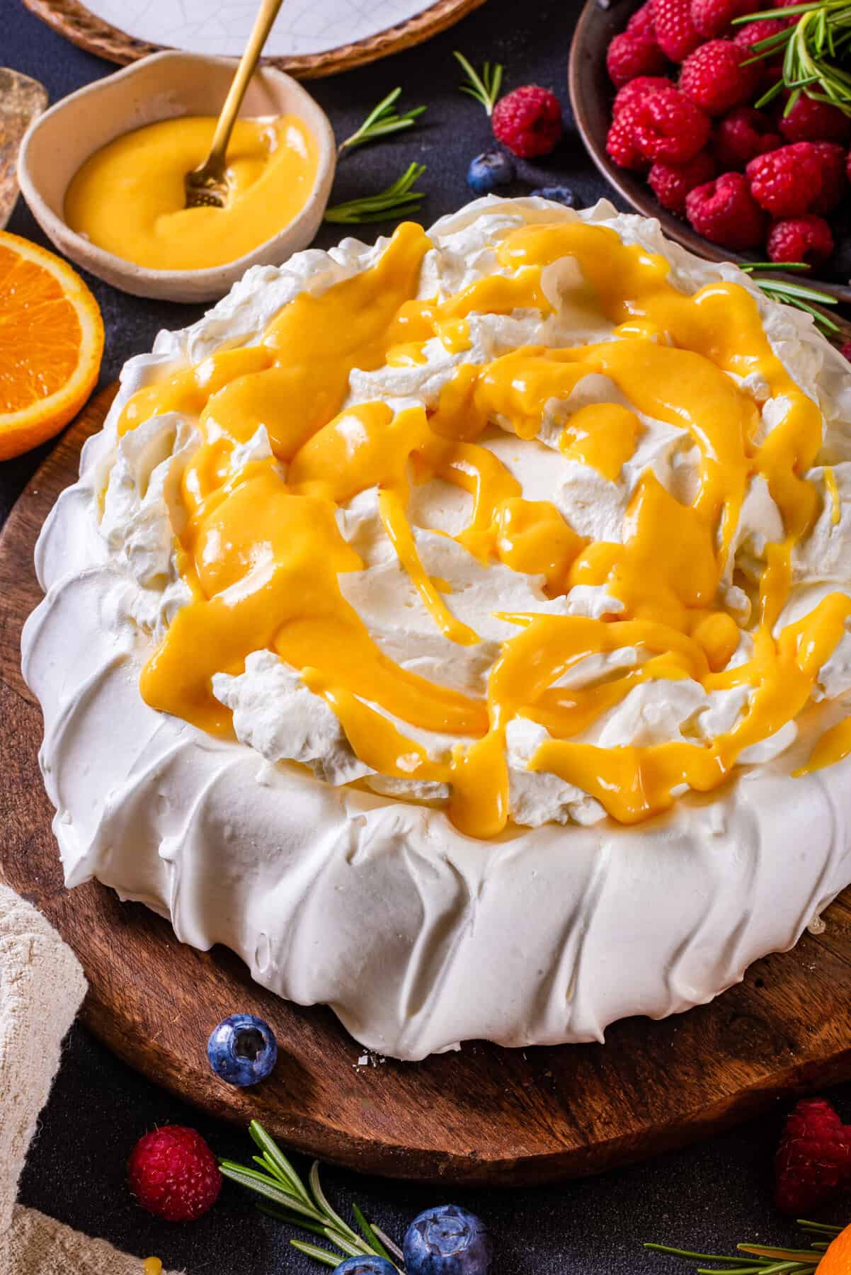 Classic Pavolva Dessert A pavlova recipe topped with whipped cream and drizzled with orange curd, surrounded by fresh blueberries, raspberries, an orange slice, and a small bowl of sauce on a wooden board.