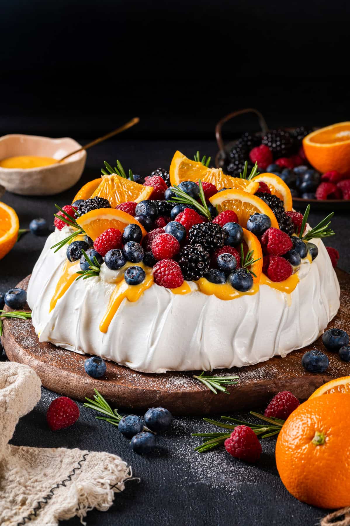 Classic Pavlova Dessert A pavlova recipe featuring a dessert topped with orange curd, orange slices, blackberries, blueberries, and raspberries, garnished with rosemary, sits on a wooden board with fresh fruit scattered around on a dark background.