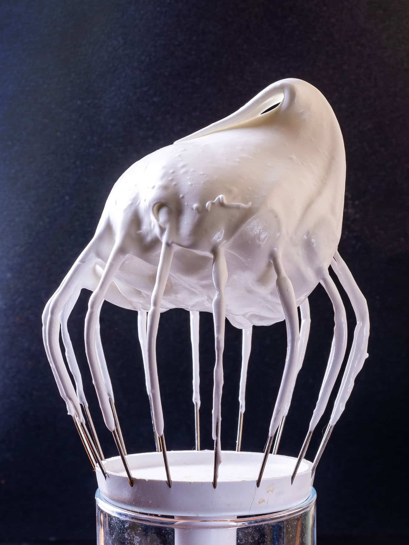 How to Make Meringue for Pavlova Dessert A whisk covered in thick, glossy, whipped meringue stands upright against a dark background.