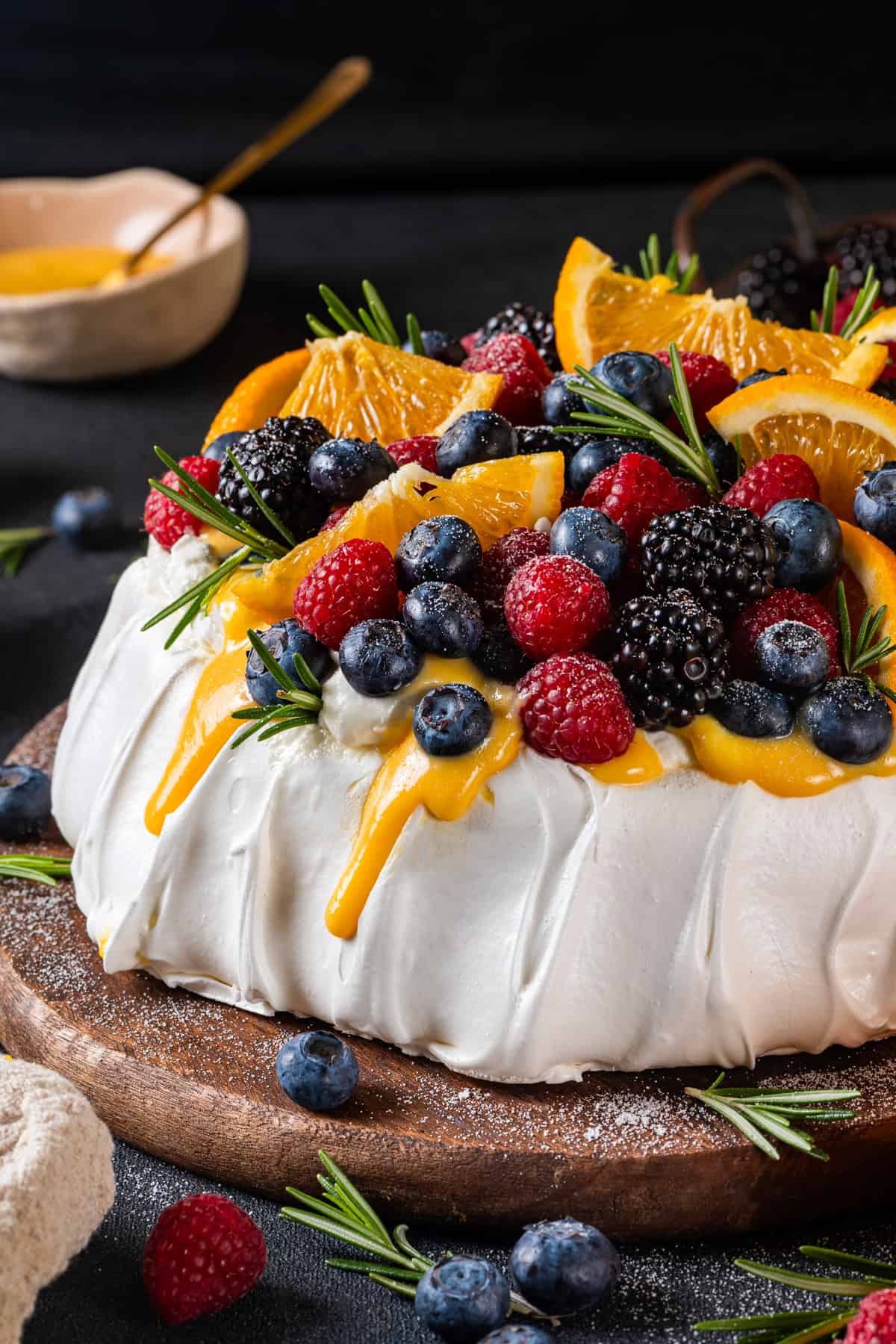 Classic Pavlova Dessert A pavlova recipe topped with whipped cream, fresh raspberries, blueberries, blackberries, orange slices, and sprigs of rosemary, with orange curd sauce drizzling down the sides, served on a wooden board.