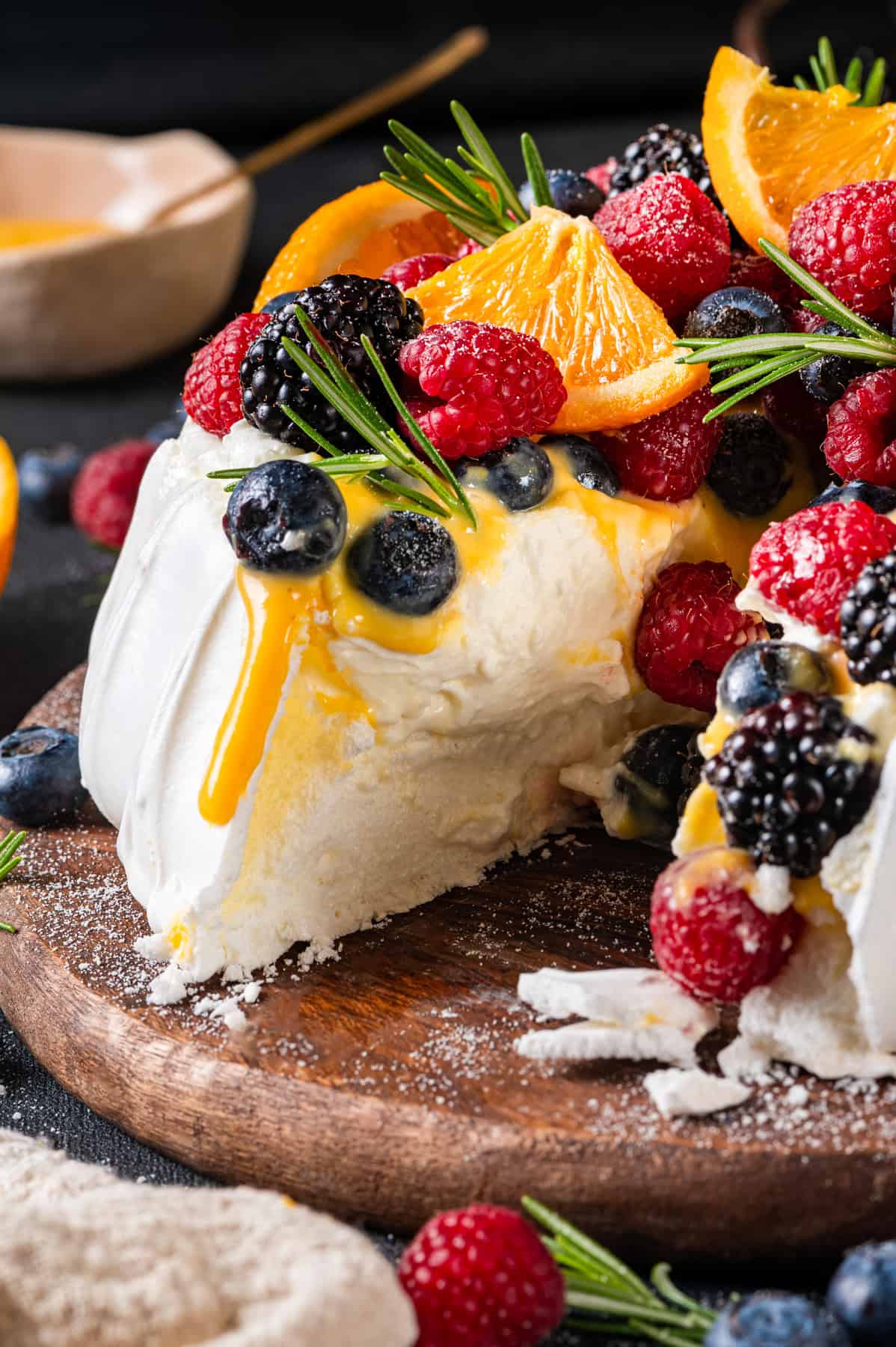 Classic Pavlova Dessert A pavlova recipe topped with fresh raspberries, blackberries, blueberries, orange slices, and rosemary. A cut reveals the creamy interior as fruit and orange curd sauce spill over the edge on a wooden board.