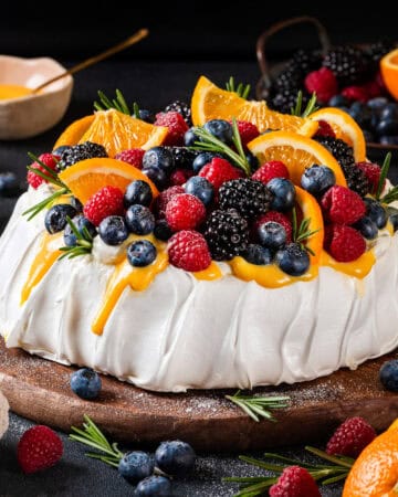A pavlova recipe topped with fresh berries, orange slices, and rosemary sits on a wooden board. Orange Curd Sauce drips down the sides, with extra fruit and orange slices scattered around on a dark background.