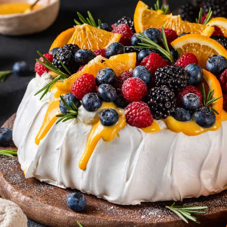 A pavlova recipe topped with whipped cream, blueberries, raspberries, blackberries, orange slices, rosemary sprigs, and drizzled with orange curd sauce, displayed on a wooden board.