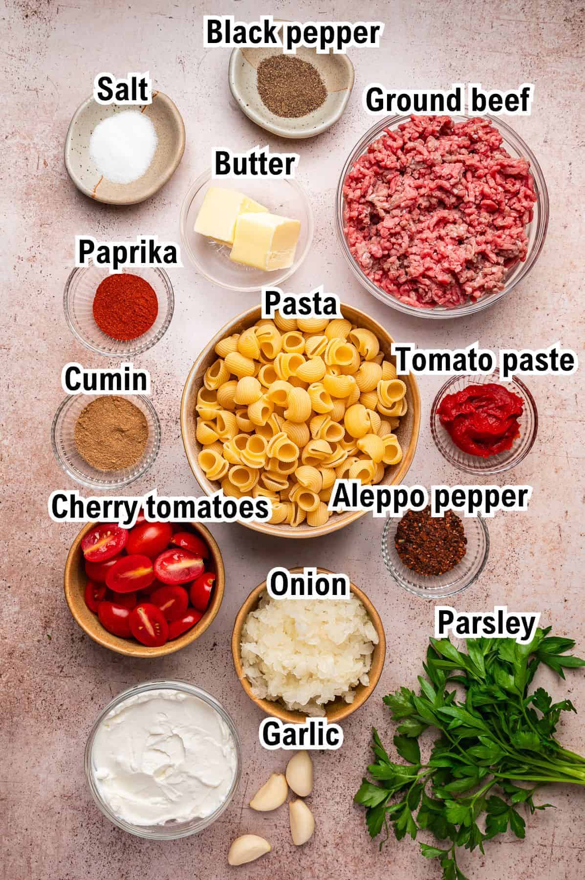 The Ingredients for Turkish Pasta Ingredients for a Turkish pasta dish arranged on a light surface, labeled: ground beef, pasta, butter, salt, black pepper, paprika, cumin, cherry tomatoes, tomato paste, Aleppo pepper, onion, garlic, parsley.