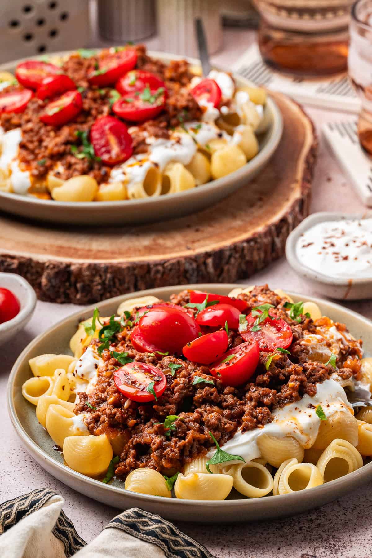 Turkish Pasta Two plates of Turkish pasta shells topped with ground meat sauce, creamy yogurt sauce, and halved cherry tomatoes, garnished with herbs. A bowl of sauce and extra cherry tomatoes sit nearby on the table.