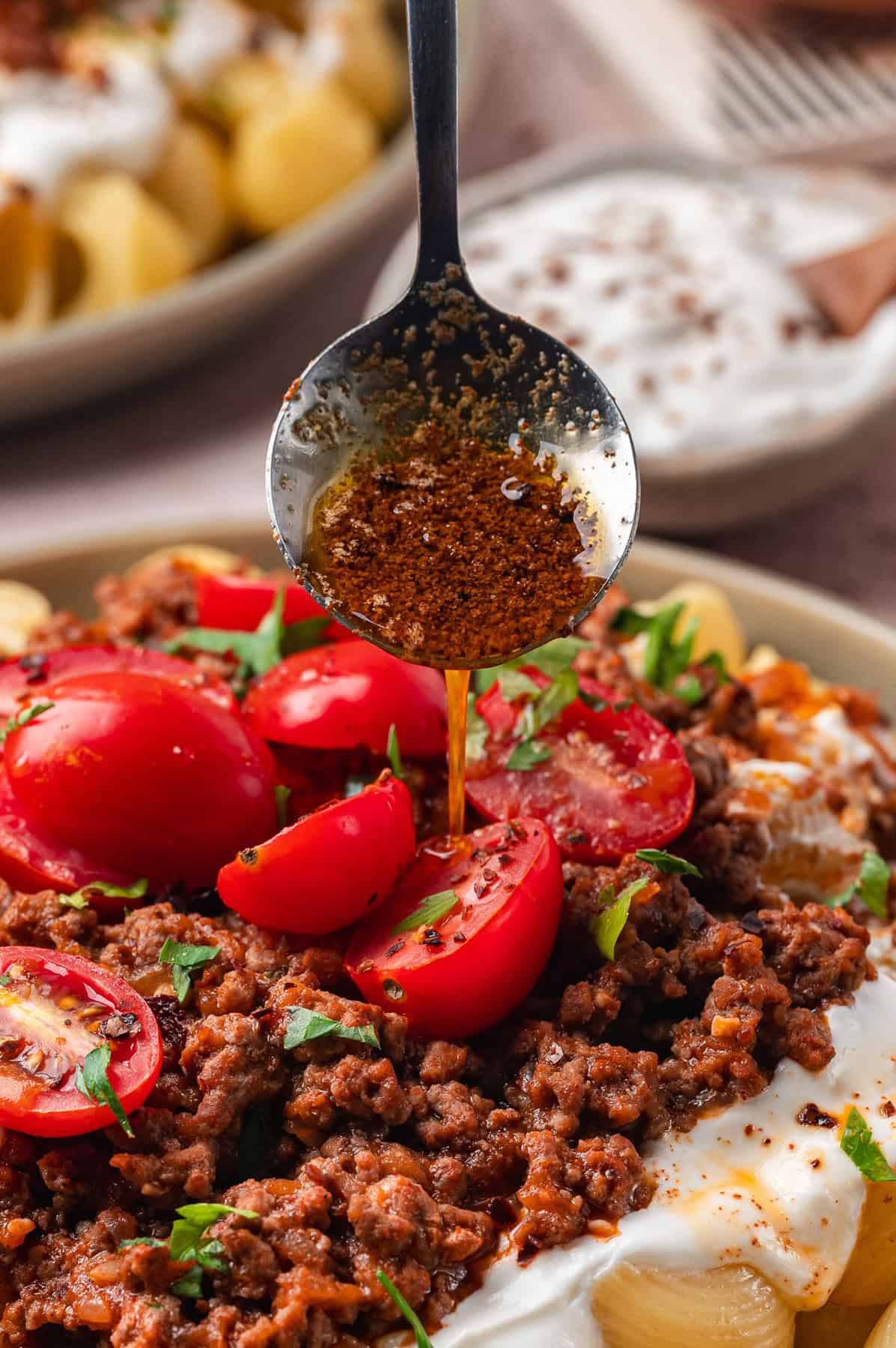 Turkish Pasta A close-up of Turkish pasta topped with ground meat, cherry tomatoes, chopped herbs, and yogurt, as a spoon drizzles spiced oil over the food.