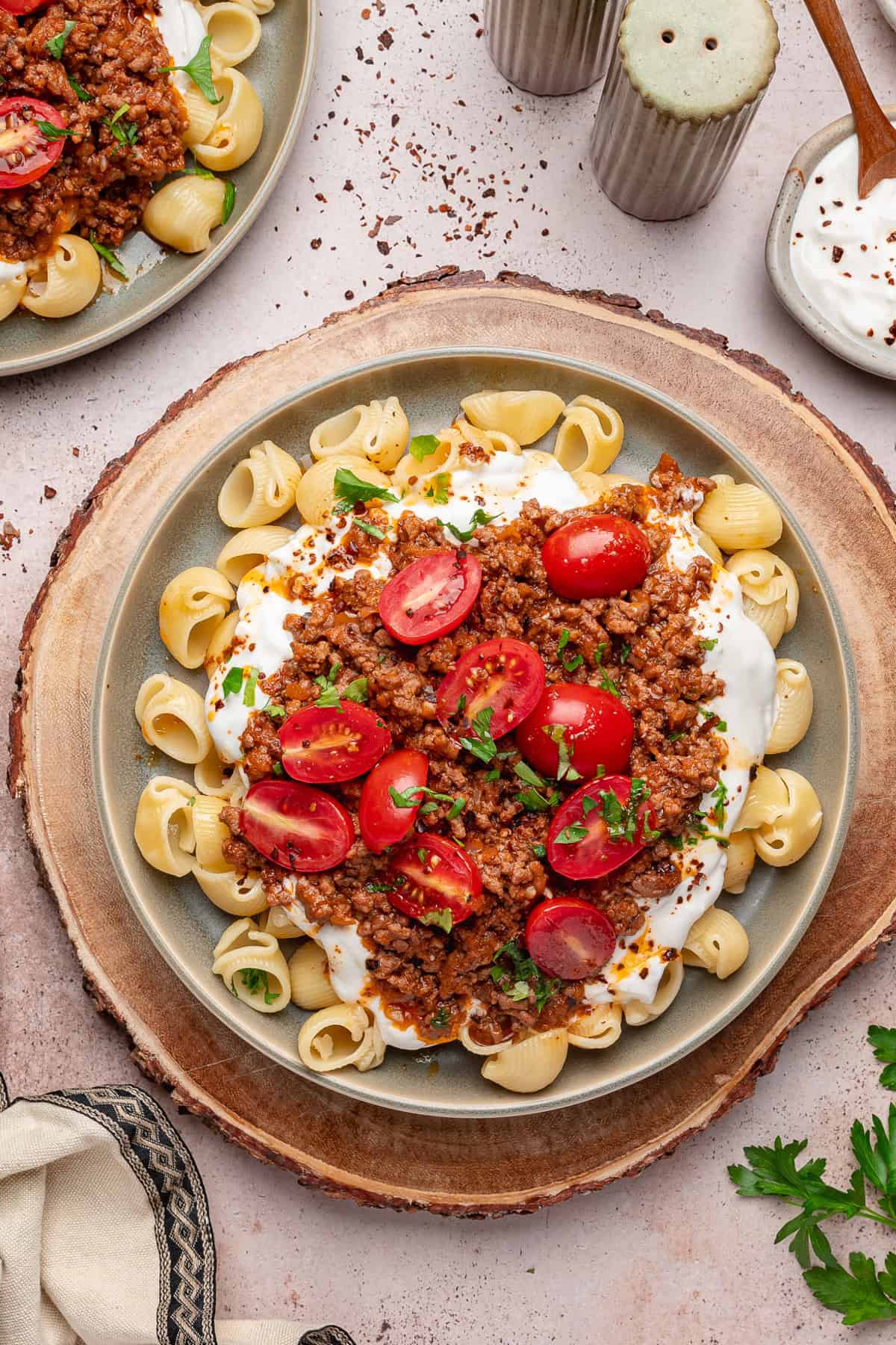 Turkish Pasta A plate of Turkish pasta shells topped with ground meat sauce, sliced cherry tomatoes, chopped herbs, and yogurt sauce, served on a wooden board with salt, pepper, and a bowl of yogurt nearby.