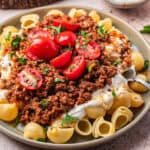 A plate of Turkish pasta shells topped with creamy white sauce, seasoned ground meat, halved cherry tomatoes, and chopped fresh parsley. A fork rests on the side of the plate.